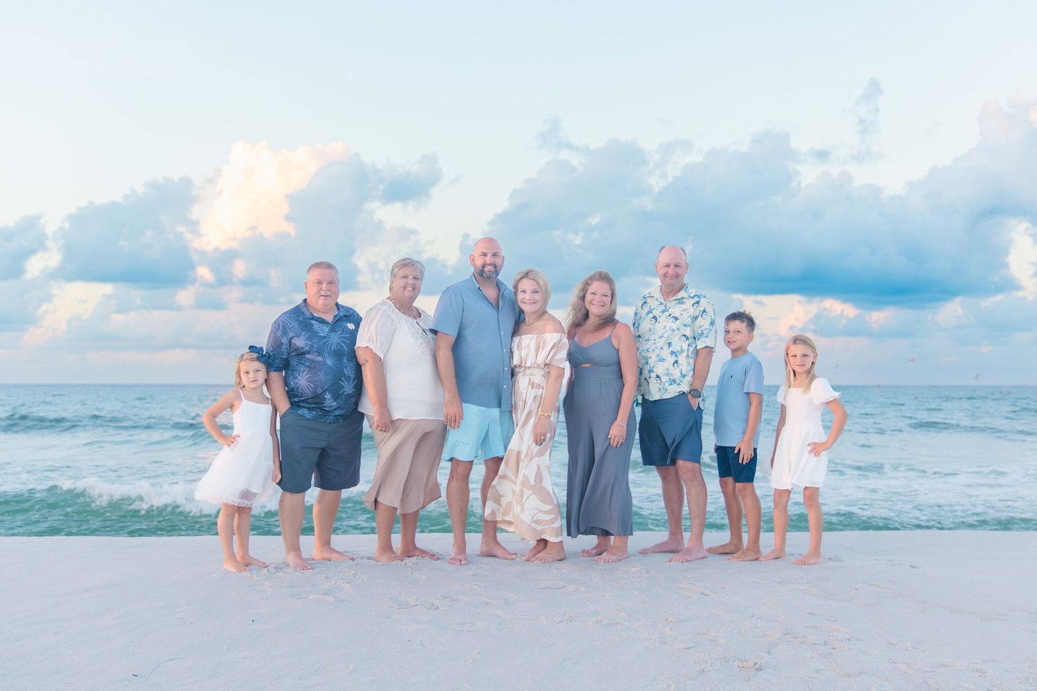 Family of nine posing together on a sandy beach at sunset with calm ocean waves and pastel clouds behind them.