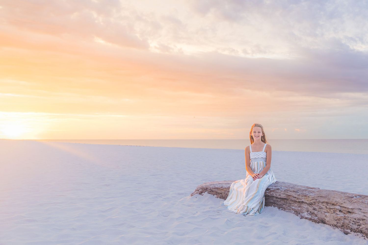 A young girl in a white dress sits on driftwood at White Sands during a pastel sunset.