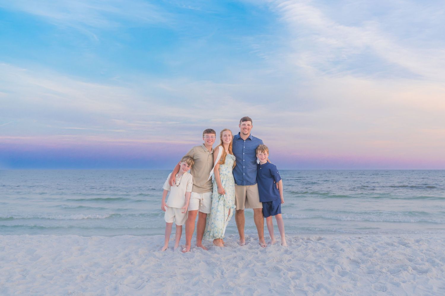 Family of five posing on a sandy beach at sunset with pastel pink and blue sky over calm ocean waters.