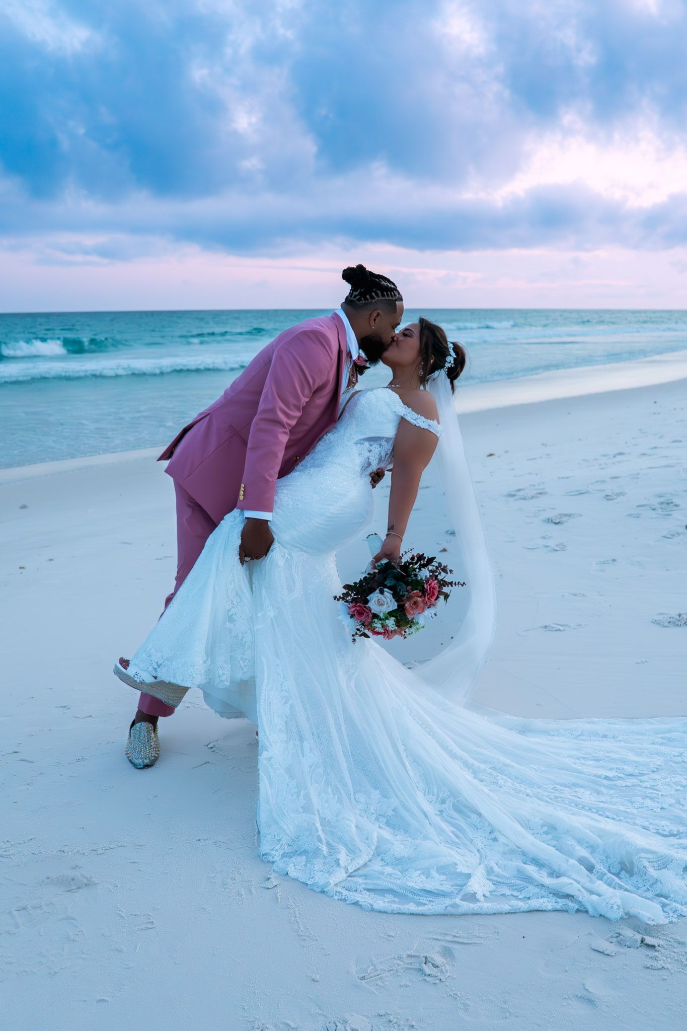 Newlywed couple sharing a romantic kiss on a white sand beach at sunset, bride in white gown, groom in pink suit.