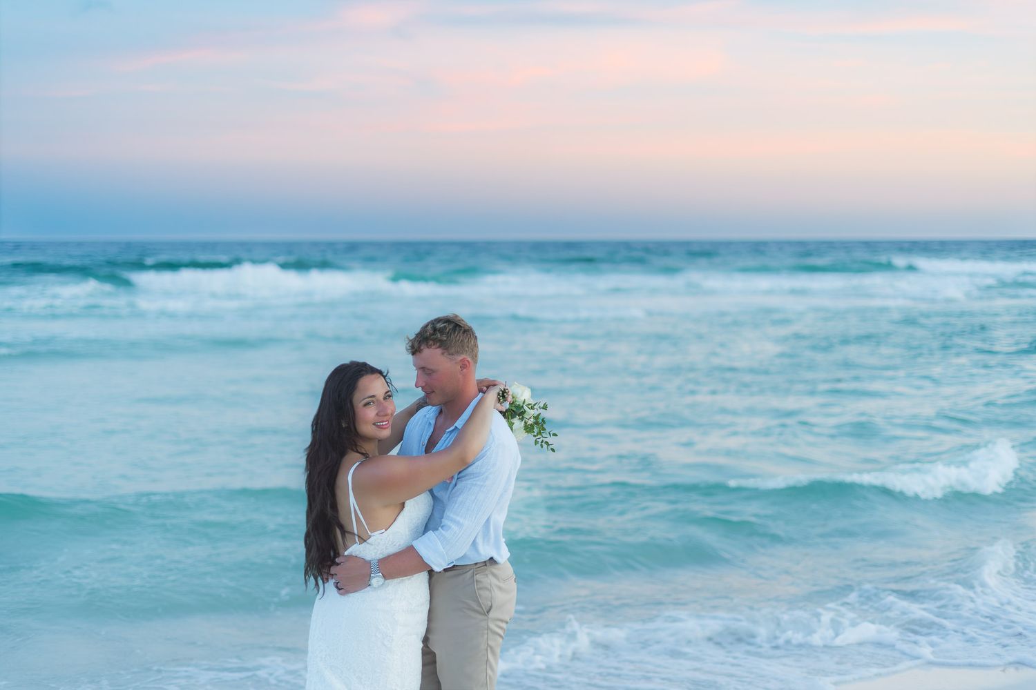Couple embracing on a beach at sunset in wedding attire, with turquoise waves and pink sky backdrop.