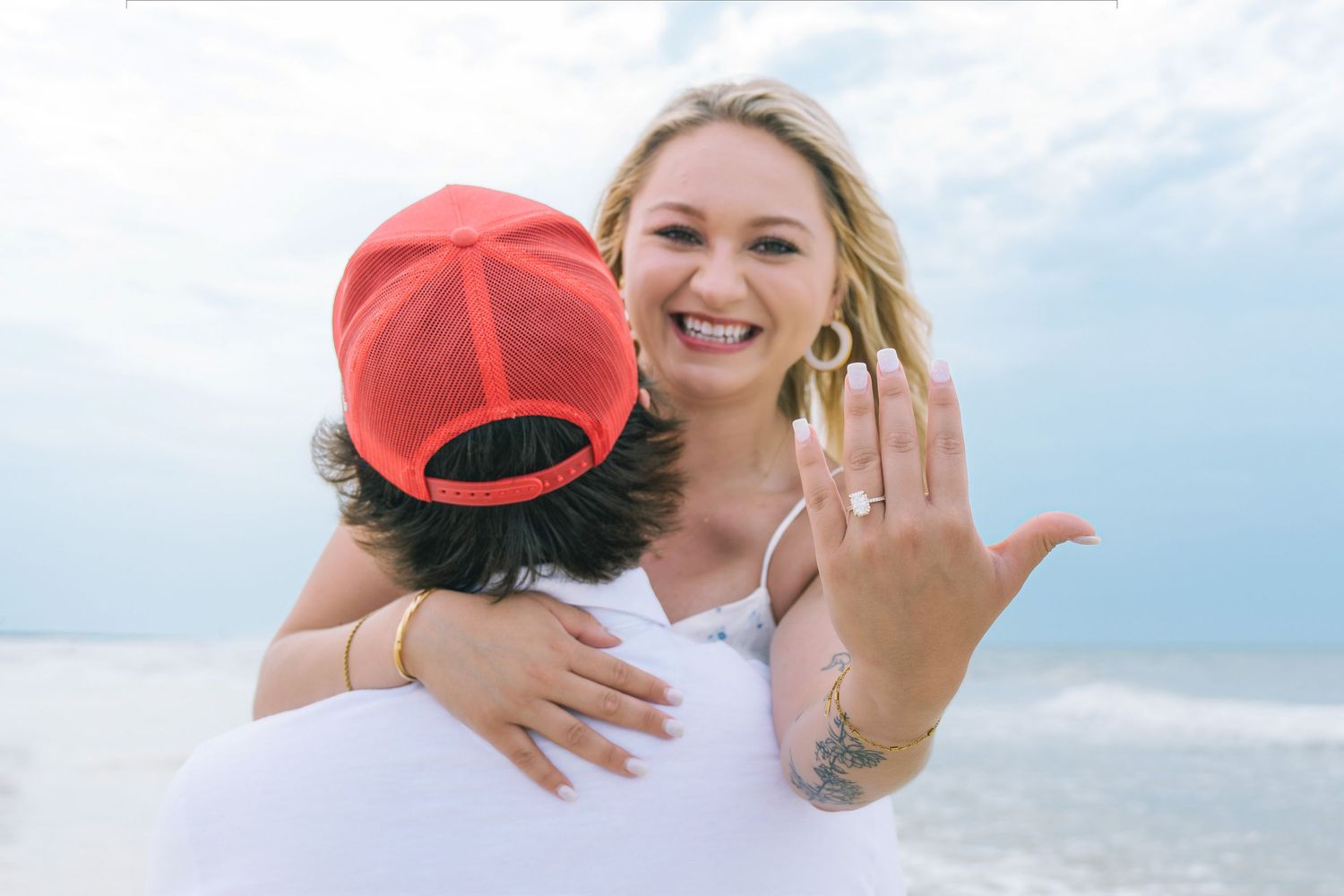 Woman in white dress showing engagement ring while embracing partner in red cap on beach.