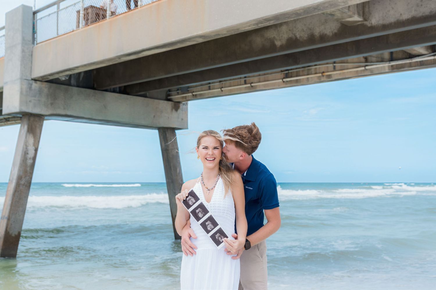 Couple on beach by pier holding ultrasound photos for pregnancy announcement, woman in white dress, man in navy polo.