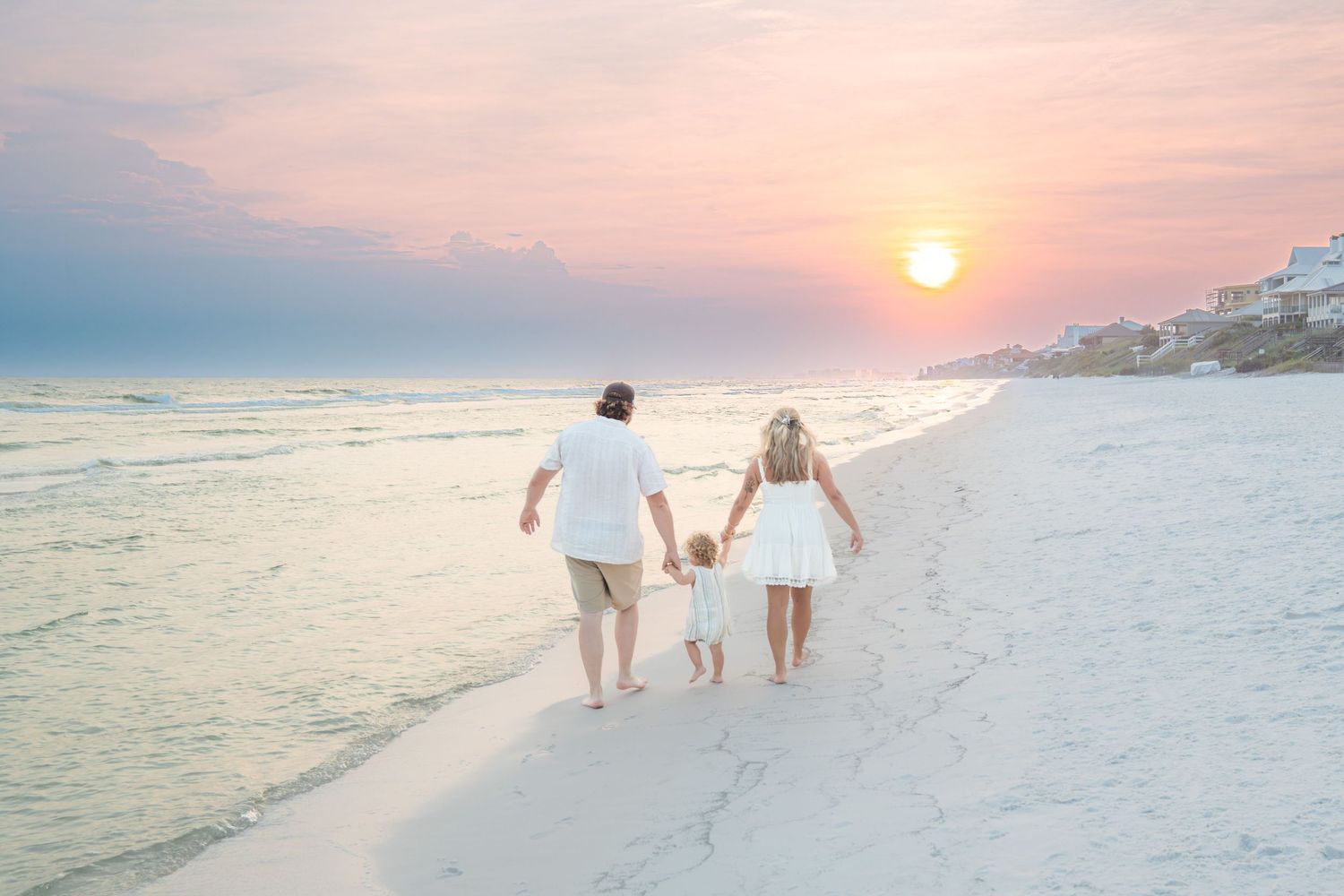 Family walking on a white sand beach at sunset, holding hands in matching white outfits with pastel sky.