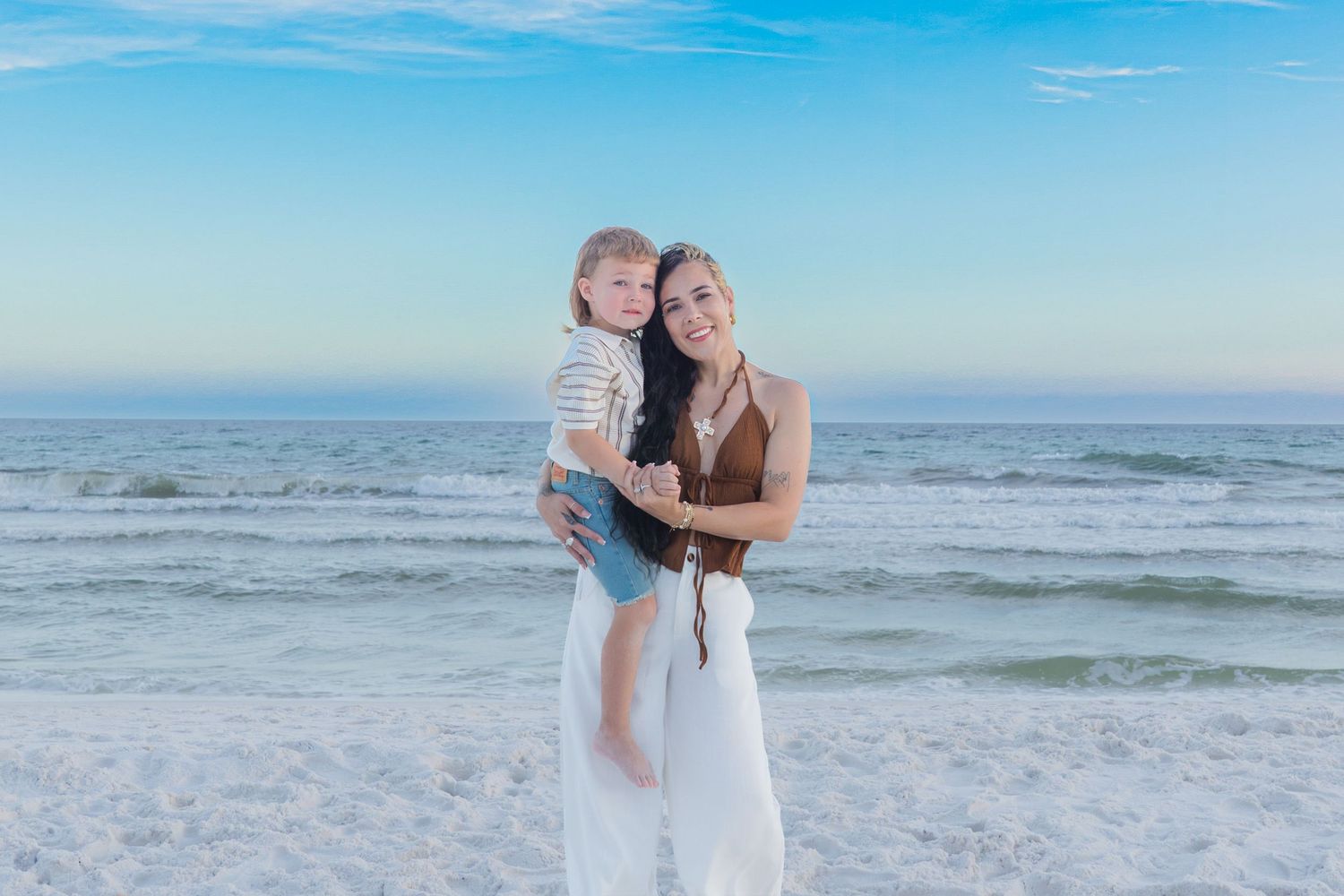 A woman in a white skirt holds a young child on a sunny beach with turquoise ocean waves in the background.
