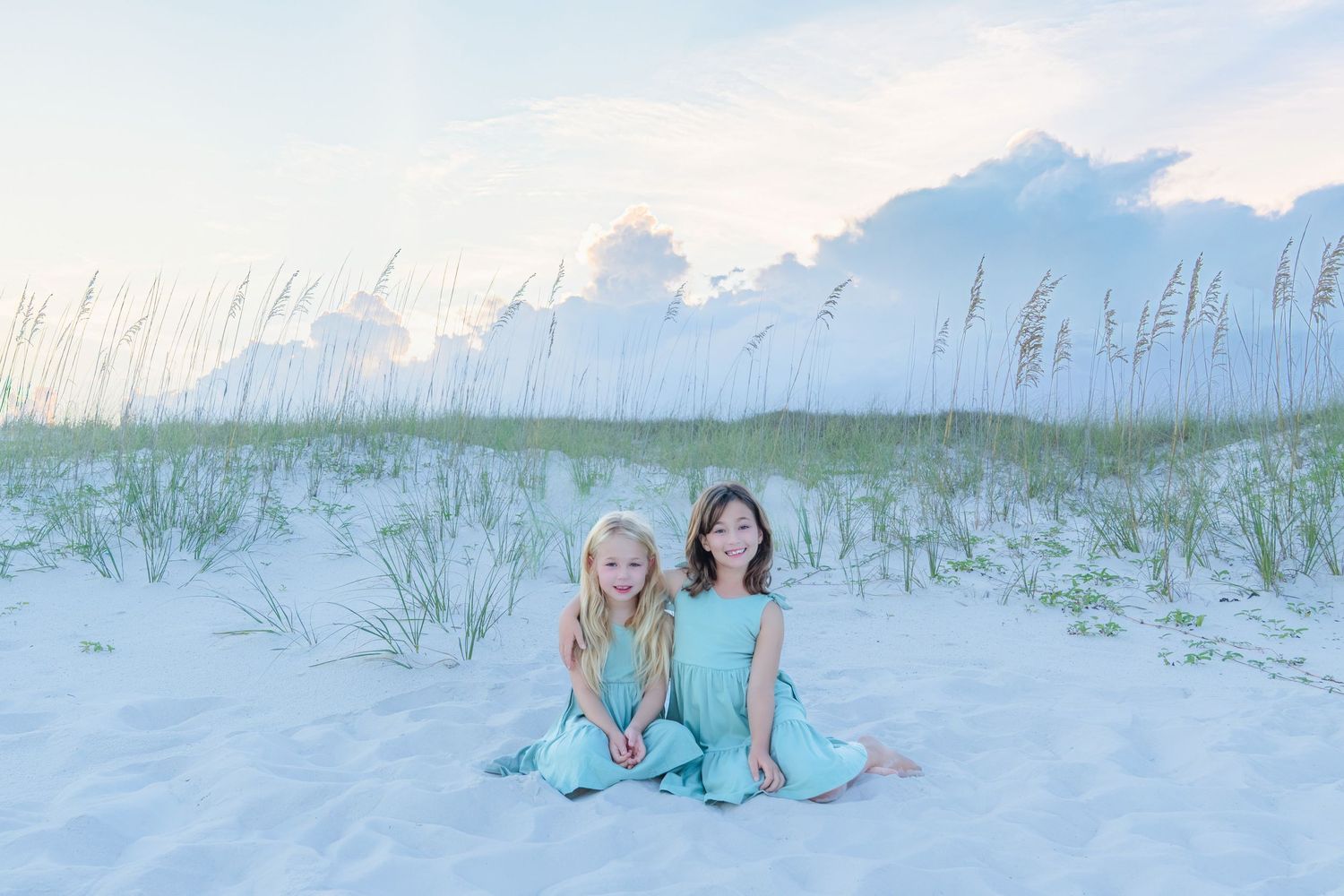 Two girls in teal outfits sit on white sand dunes with sea oats and pastel sky at a beach.