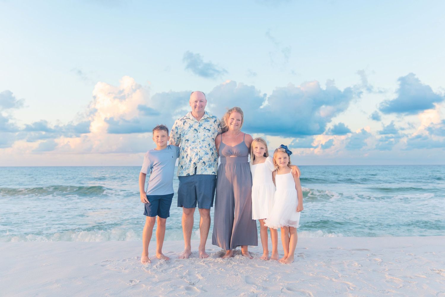 Family of five posing together on a sandy beach at sunset with calm ocean waves and pastel sky behind them.