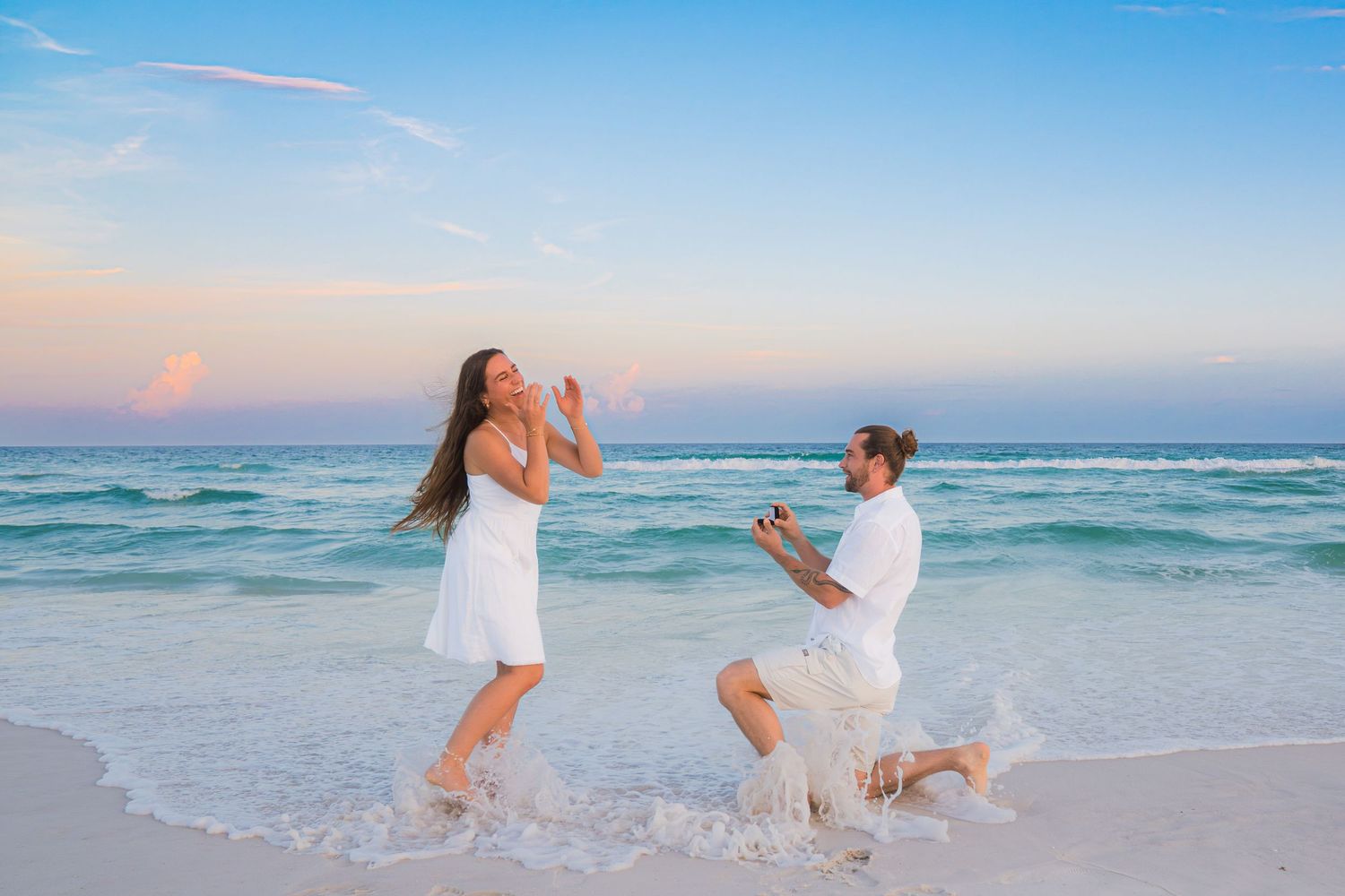 Man proposes to woman on a beautiful beach at sunset, both wearing white outfits with turquoise ocean waves.