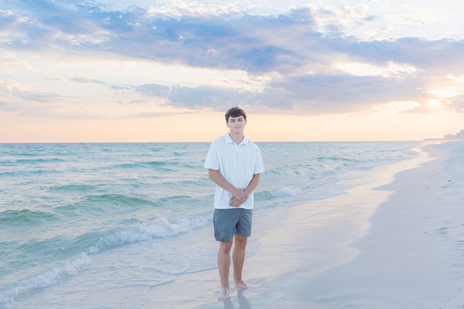 Young man in white shirt and gray shorts standing barefoot on a sandy beach at sunset with pastel sky.