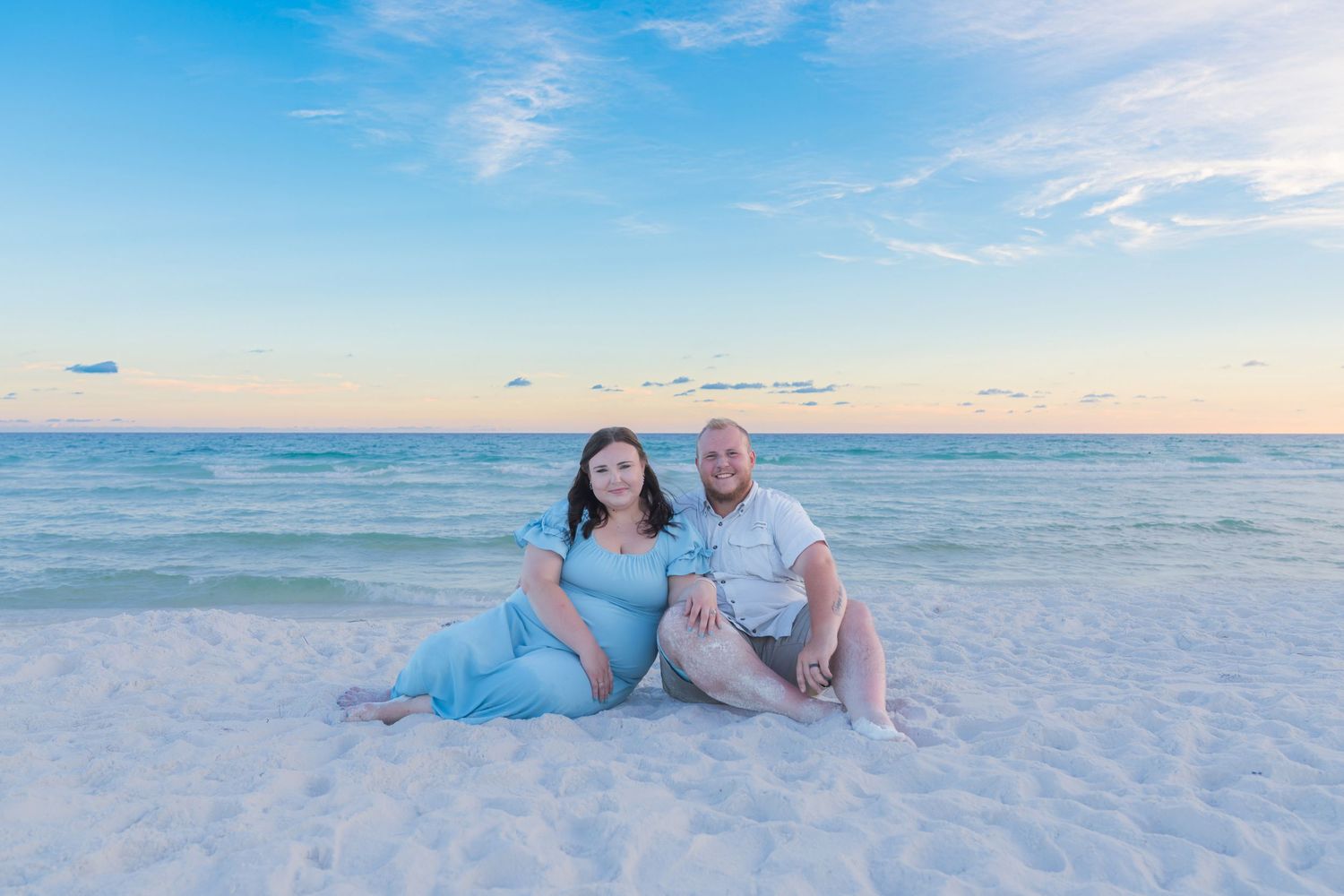 Pregnant couple sitting on white sand beach at sunset, woman in blue maternity dress, calm turquoise ocean backdrop.