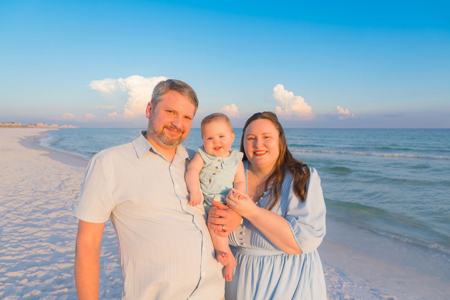 A happy family of three poses on a white sand beach at sunset, parents holding their smiling baby by turquoise waters.