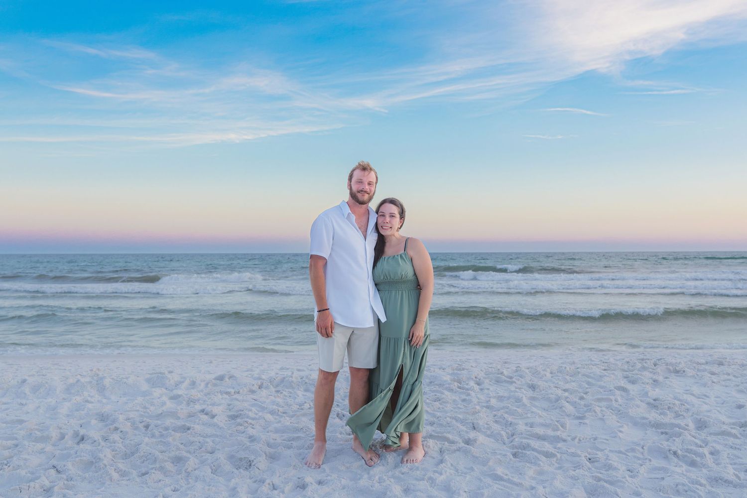 Couple smiling together on a white sand beach at sunset, man in white shirt and woman in green dress.