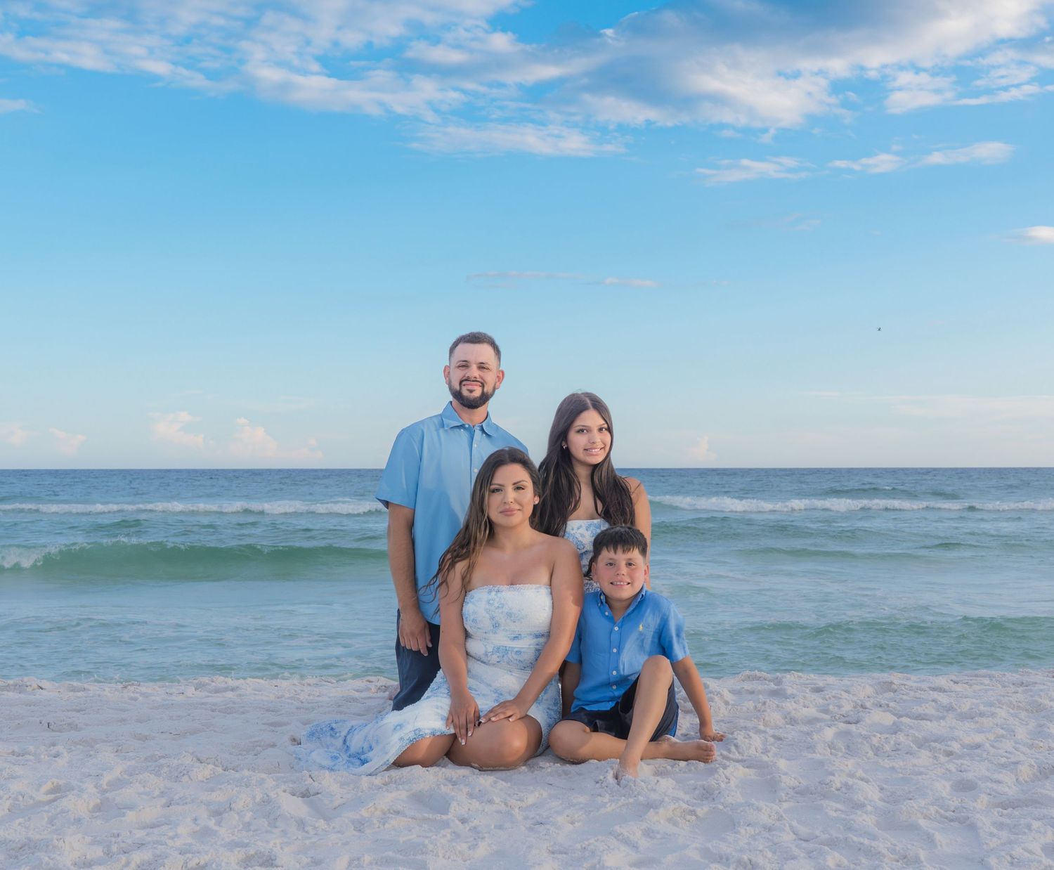 Family of four posing on a white sand beach with turquoise ocean waves and blue sky backdrop.