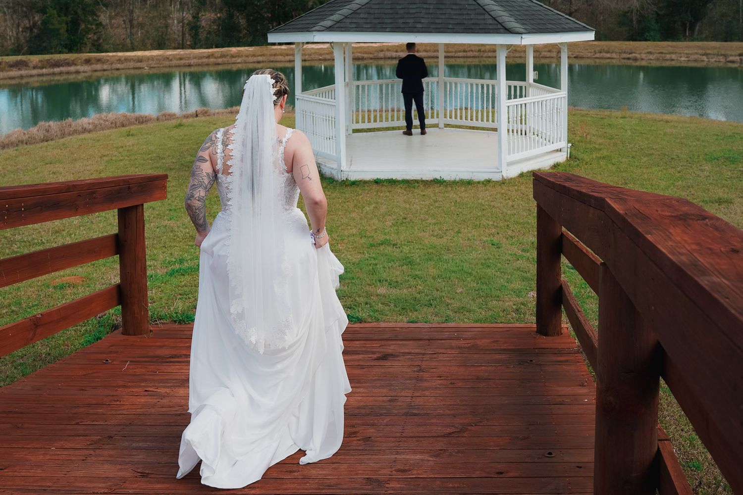 Bride in white gown and veil walks toward groom standing at gazebo by pond for wedding first look.