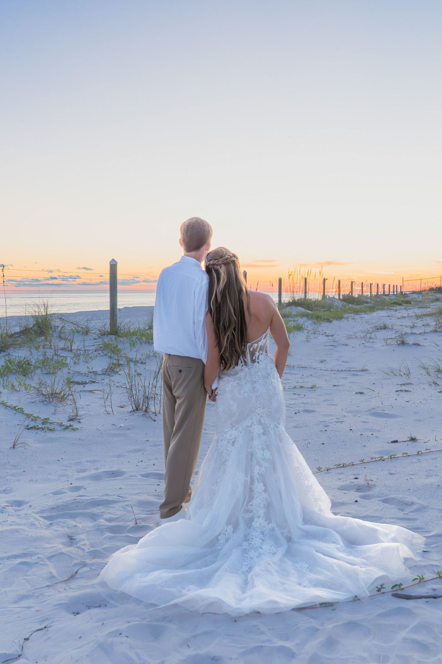 Bride and groom stand hand in hand on a sandy beach at sunset, her lace gown trailing elegantly behind them.