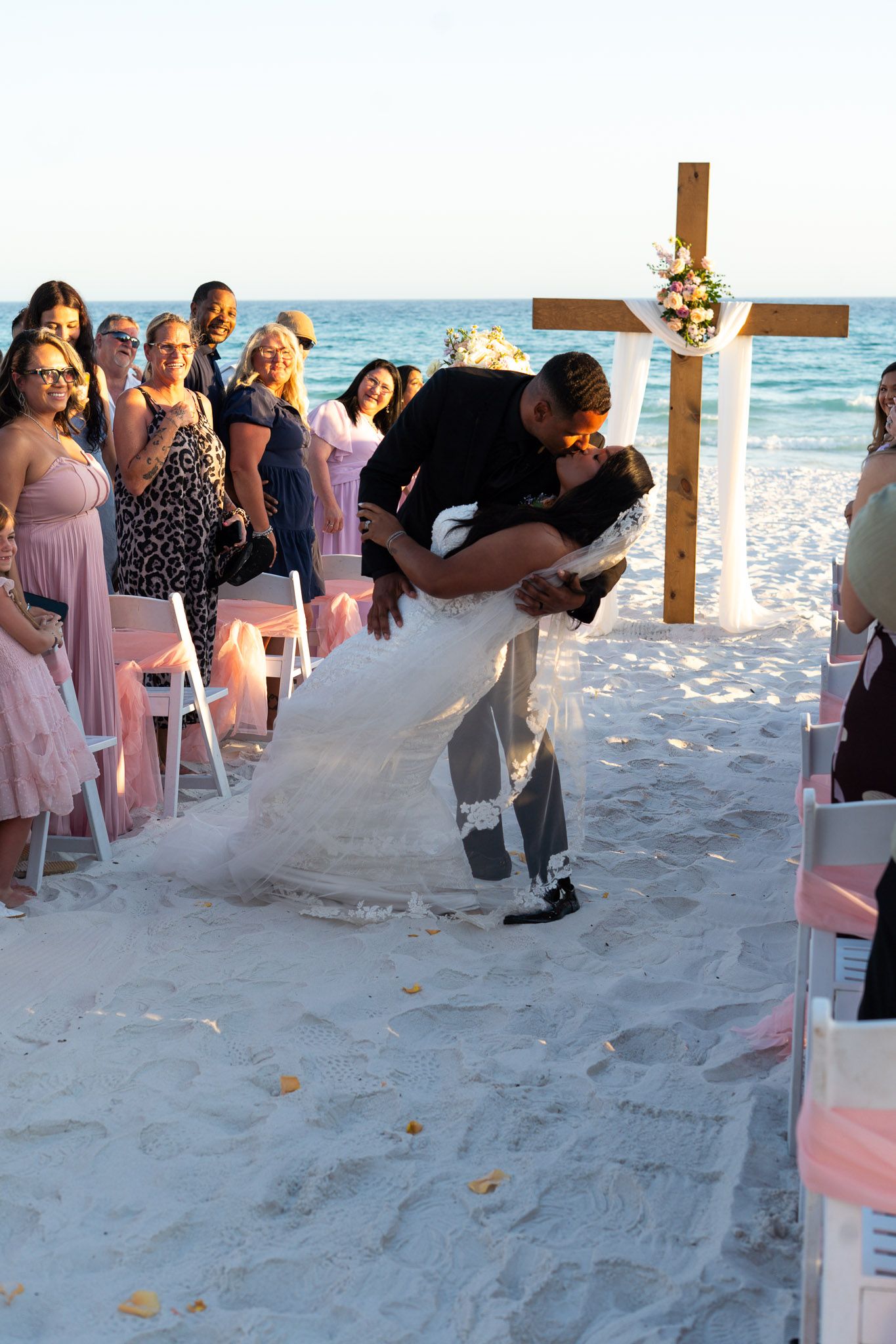 Groom dipping bride for a romantic kiss on sandy beach aisle after outdoor wedding ceremony at sunset.
