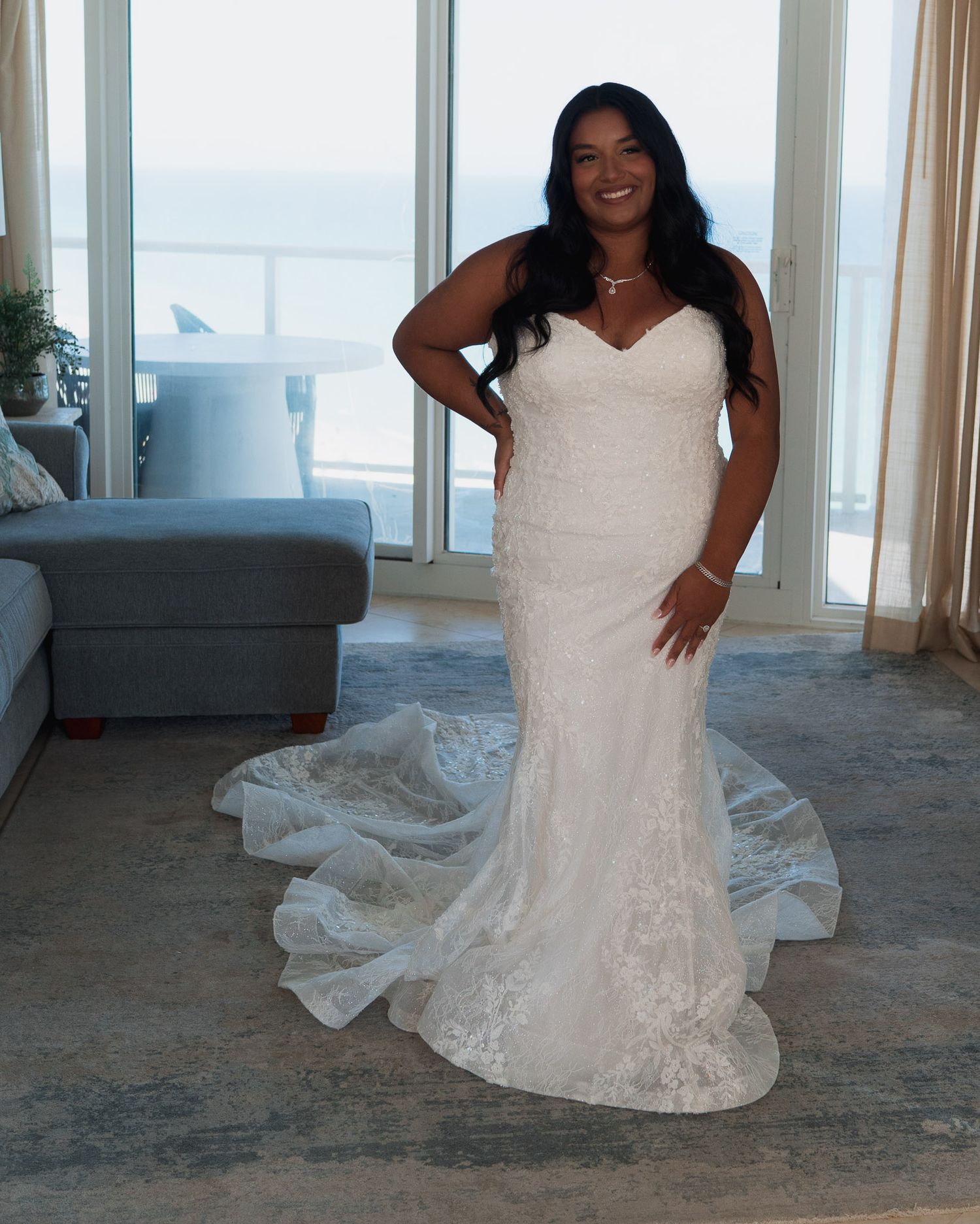 Bride in strapless lace mermaid wedding gown with long train posing by ocean view window.