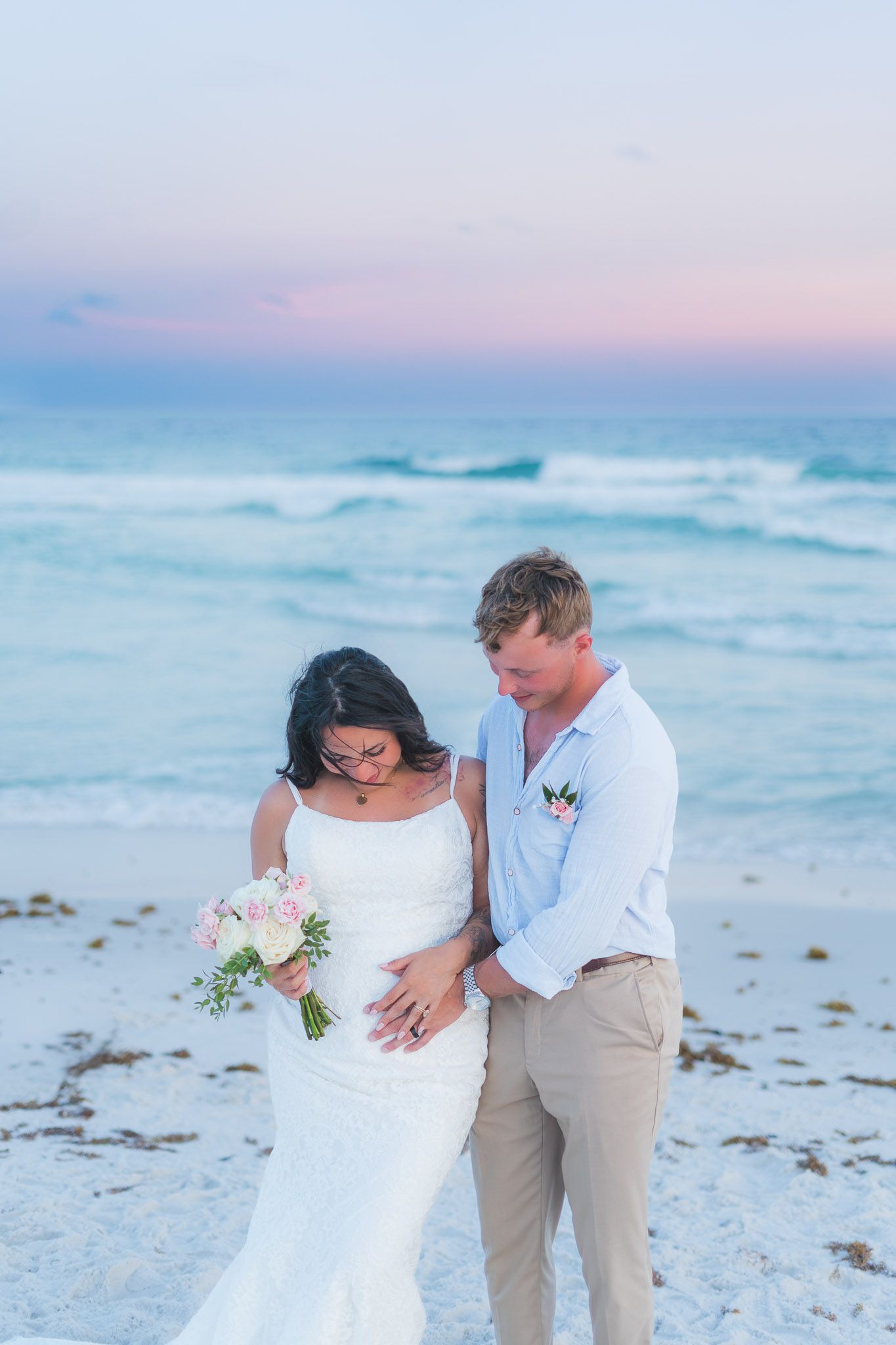 Newlyweds embracing on a beach at sunset, bride holding a pink bouquet.