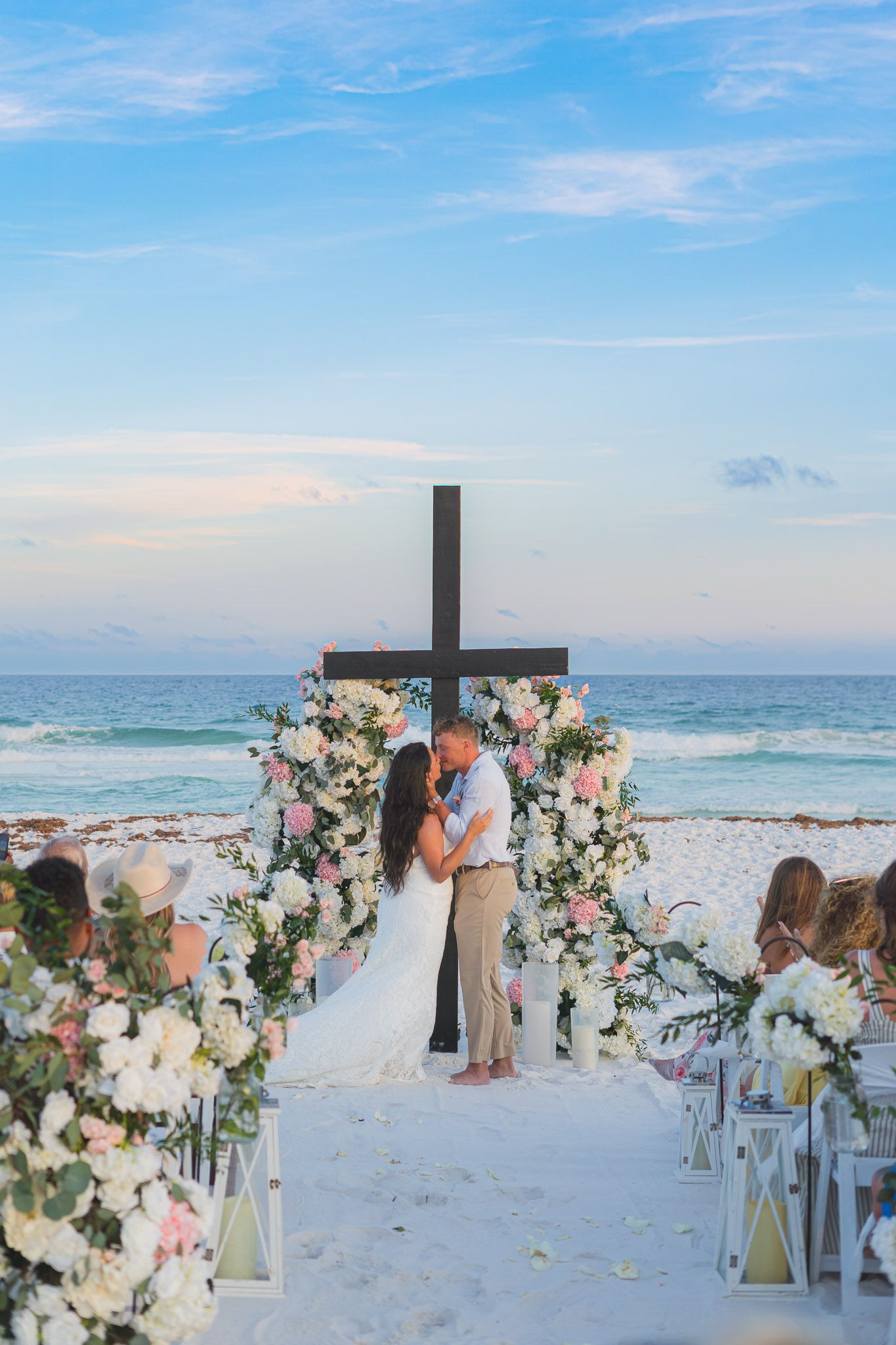 Couple kissing at beach wedding ceremony with floral arch, wooden cross, and ocean backdrop at sunset.