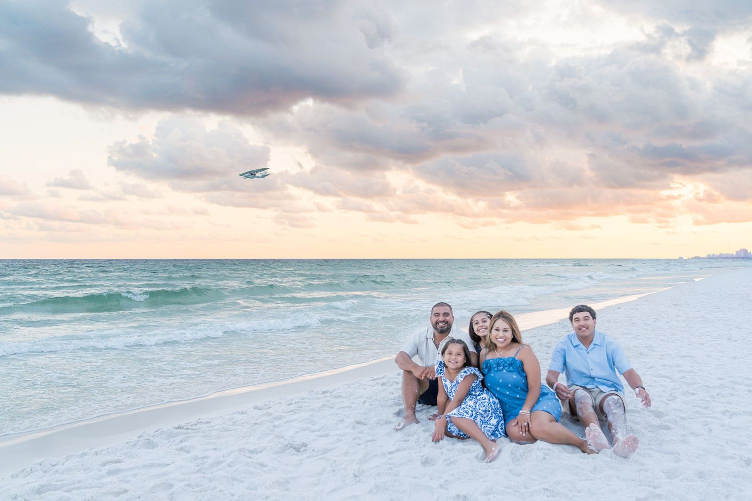 Family of five posing on white sand beach at sunset with turquoise water and colorful sky in background.