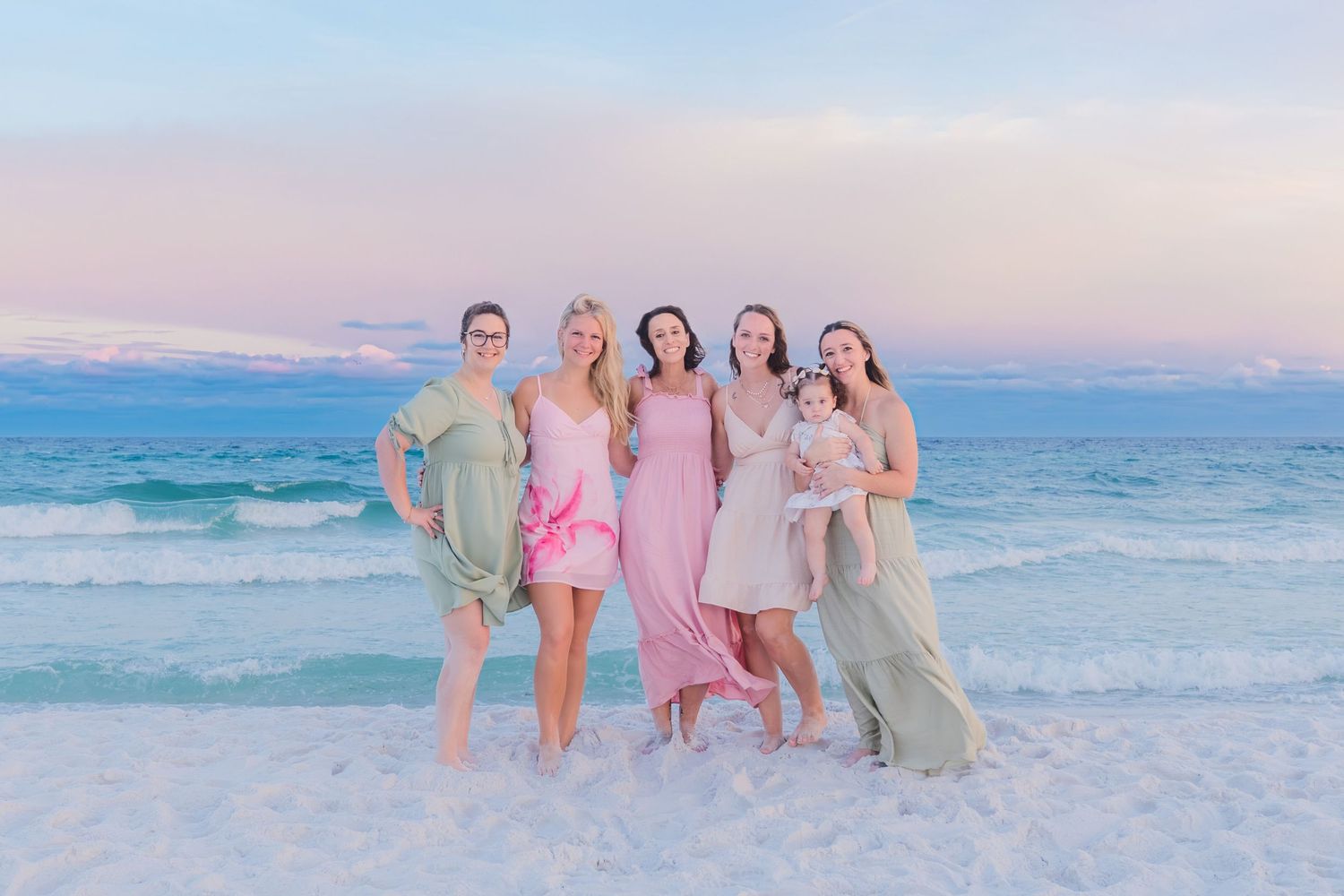 Five women in pastel dresses posing together on a beach at sunset with turquoise waves behind them.