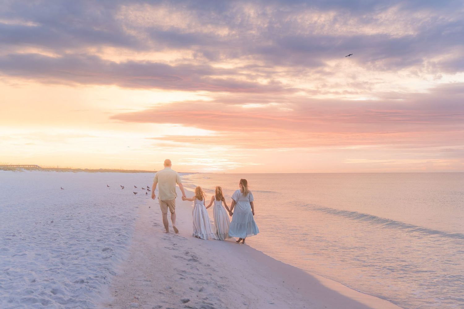 Family of four walking hand-in-hand on a white sand beach at sunset with pastel pink and purple skies.