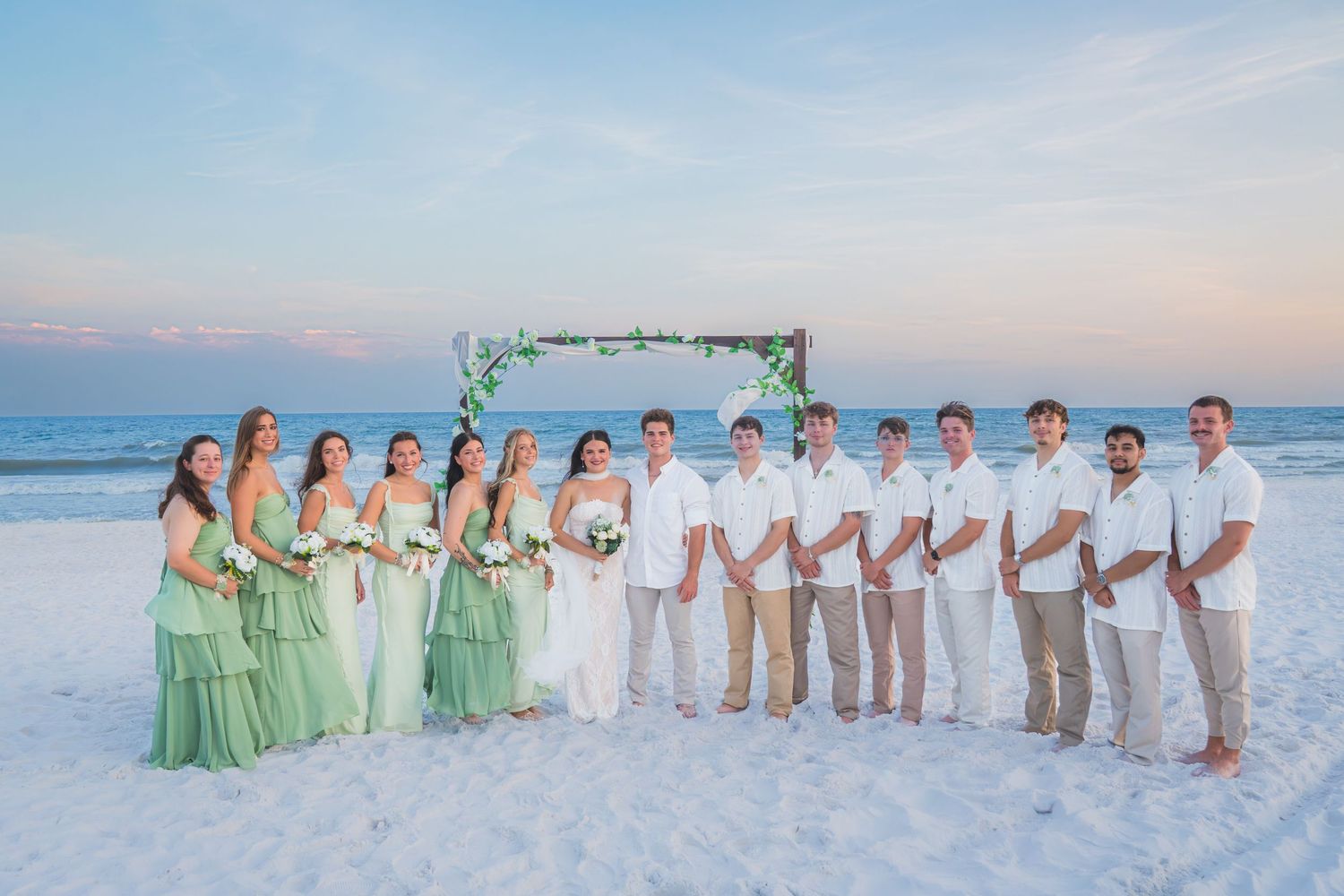 Wedding party in green and white attire posing on beach at sunset with floral arch backdrop.