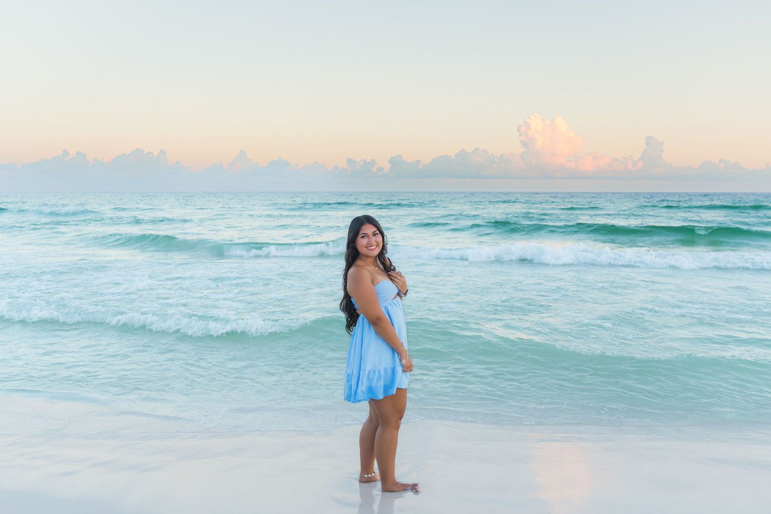 Young woman in blue dress smiling on a white sand beach with turquoise waves and pastel sunset sky.