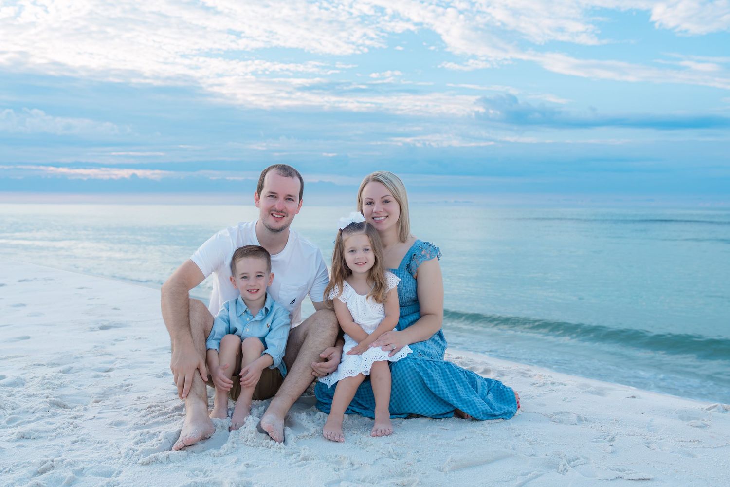 Family of four smiling together on a white sand beach at sunset with calm turquoise ocean in the background.