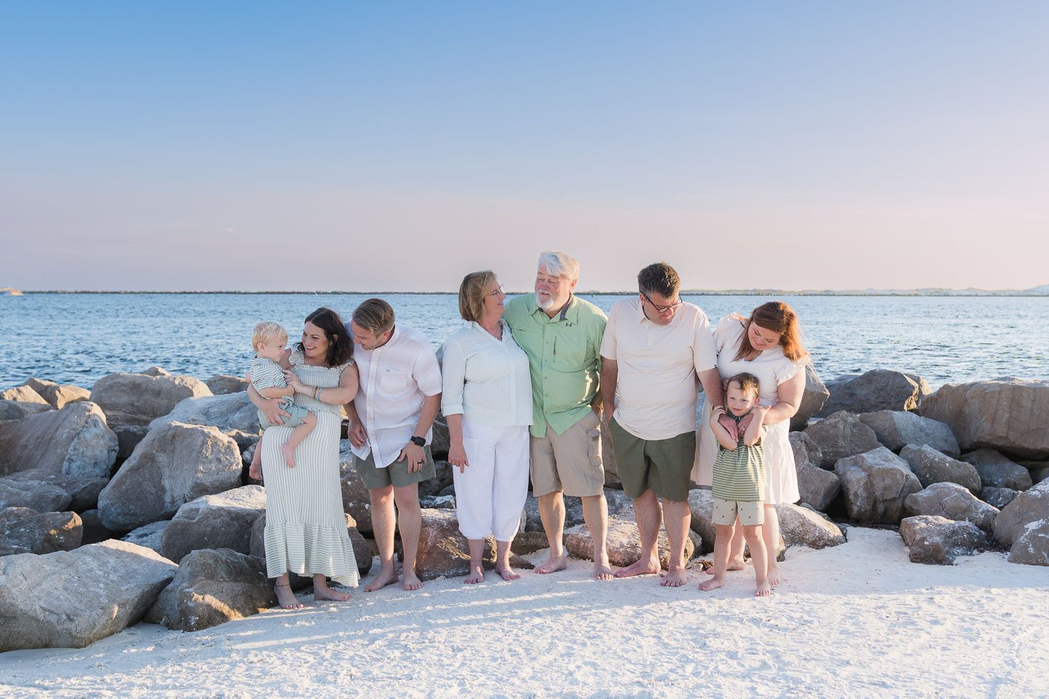 Multi-generational family posing together on a sandy beach near rocky jetty at sunset in casual white and green outfits.