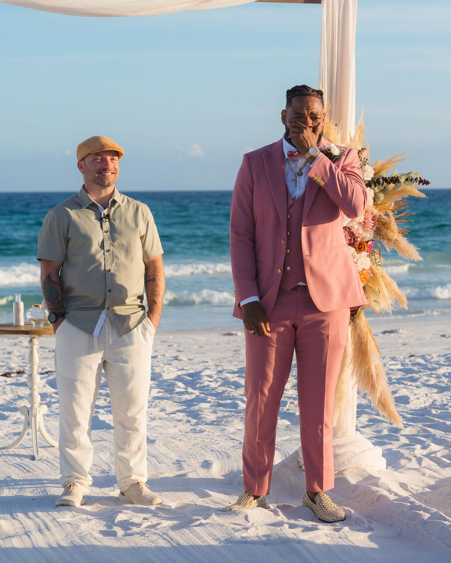 Two men stand on a sandy beach wedding ceremony, one in a pink suit and one in casual beige attire, with ocean waves behind.