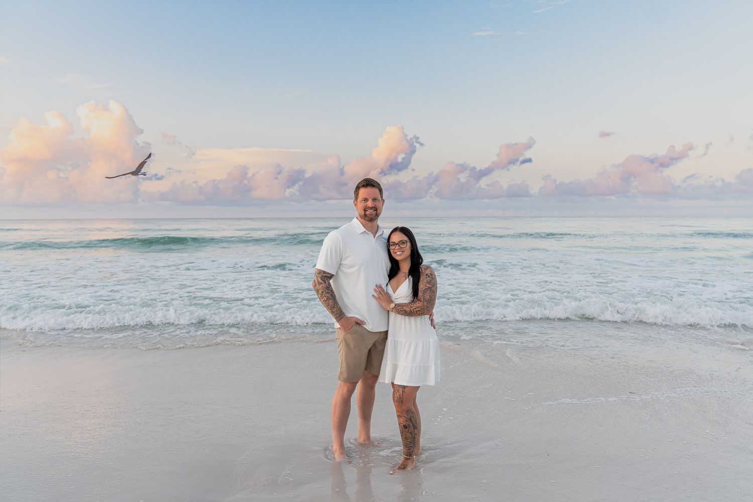 Couple smiling together on a sandy beach at sunset with turquoise waves and pastel clouds behind them.