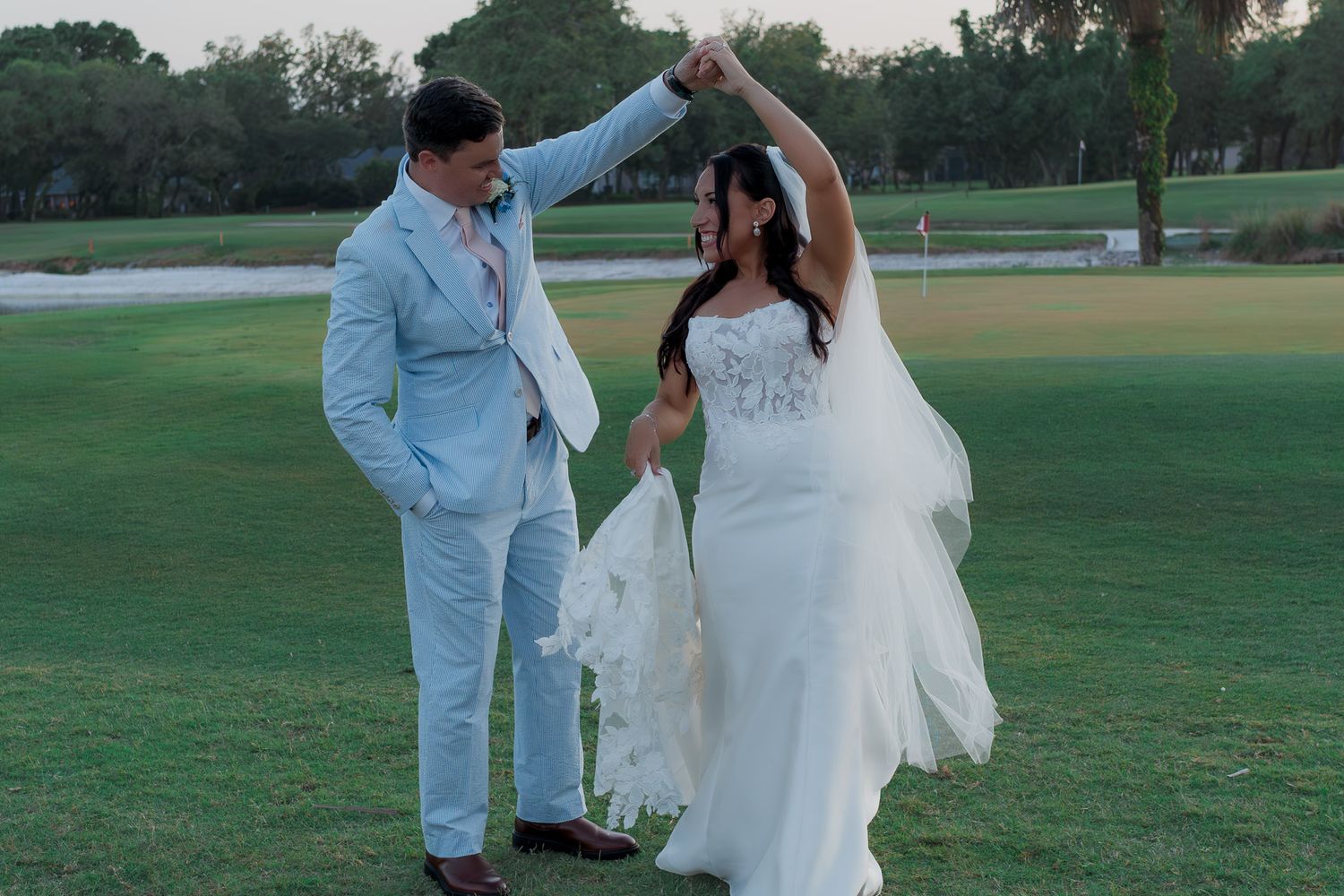 Groom in light blue suit twirls bride in white wedding dress on lush golf course at dusk.