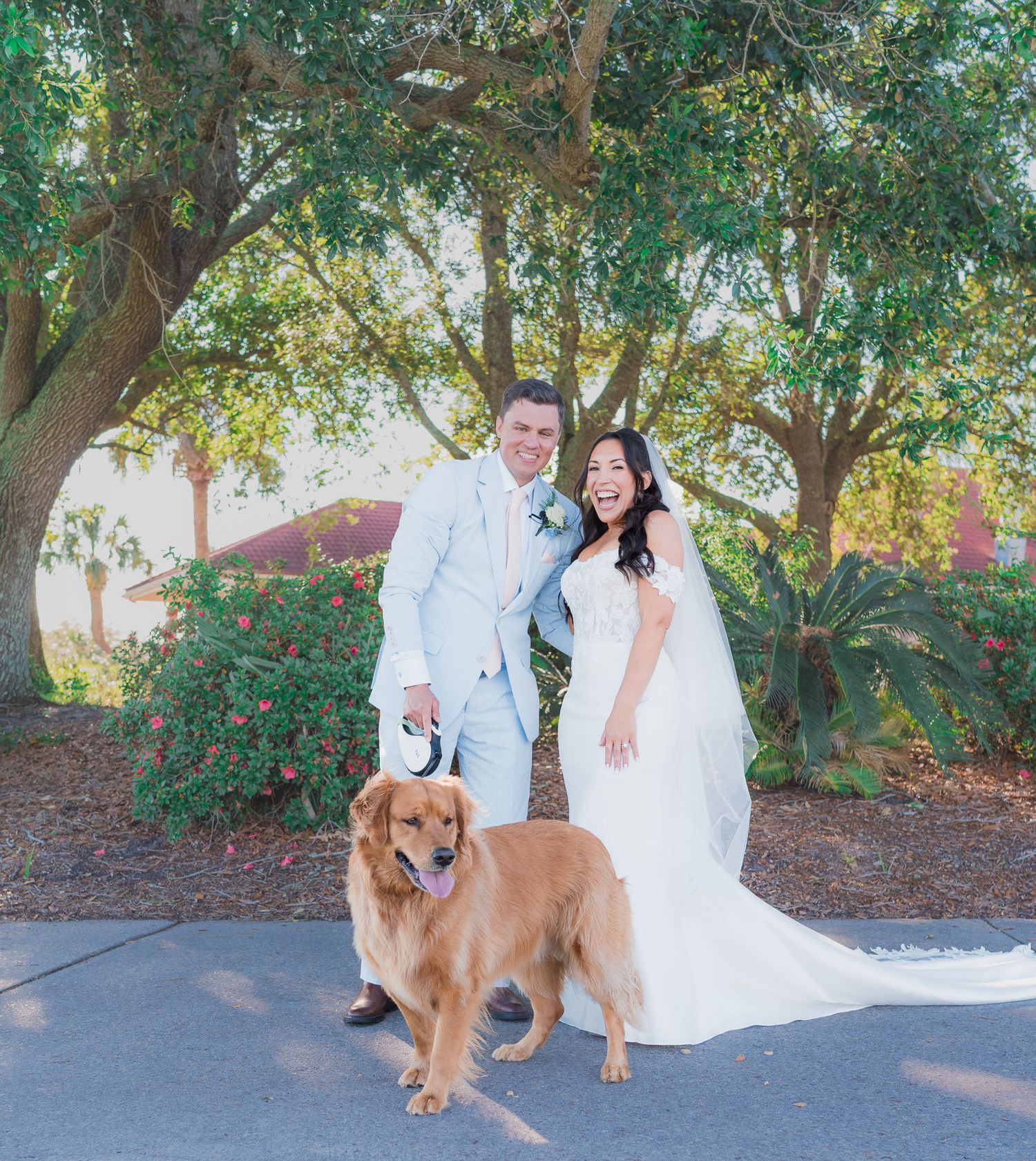 Bride and groom smiling with their golden retriever dog outdoors on their wedding day.