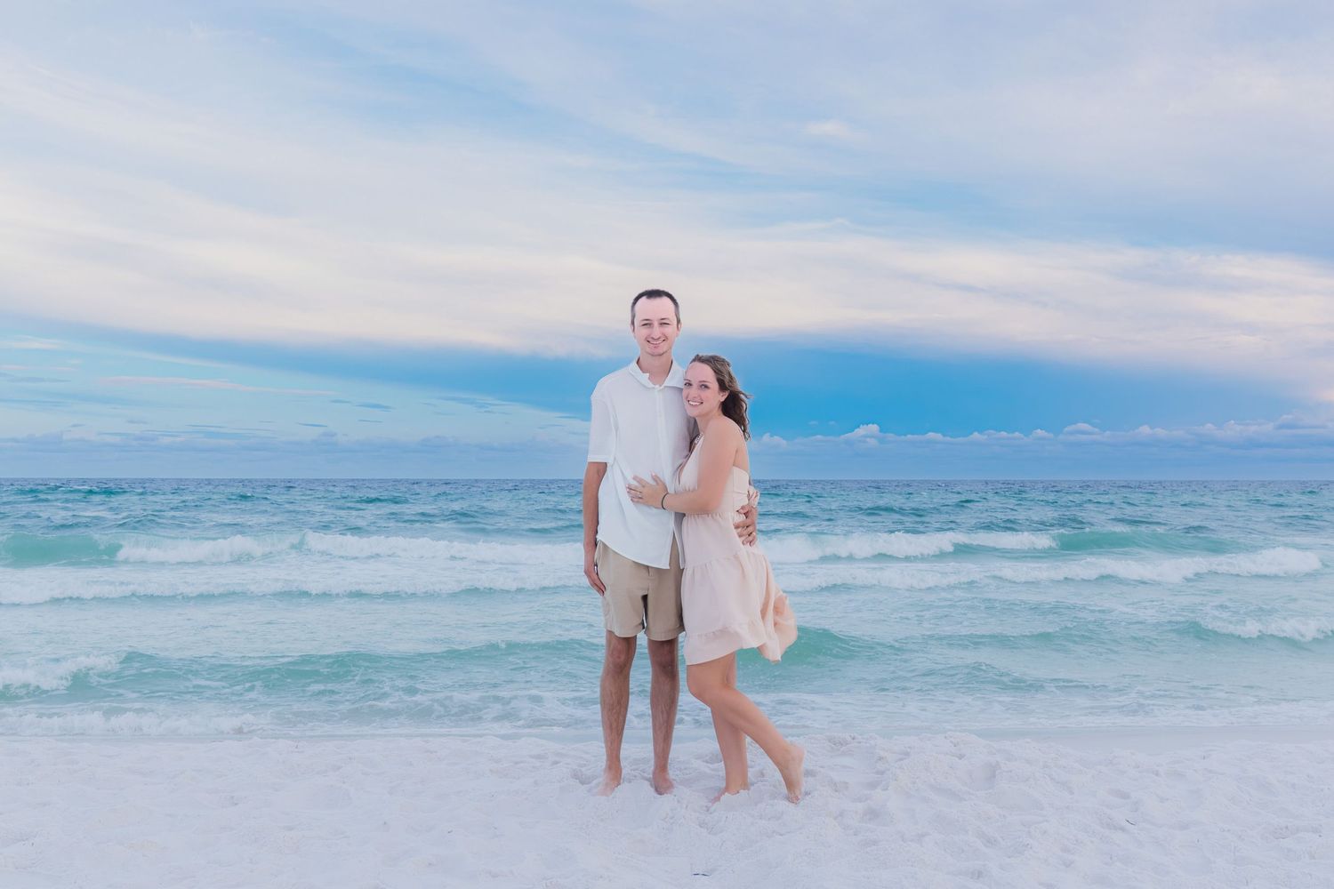 Couple smiling together on a sandy beach with turquoise waves and pastel sunset sky behind them.