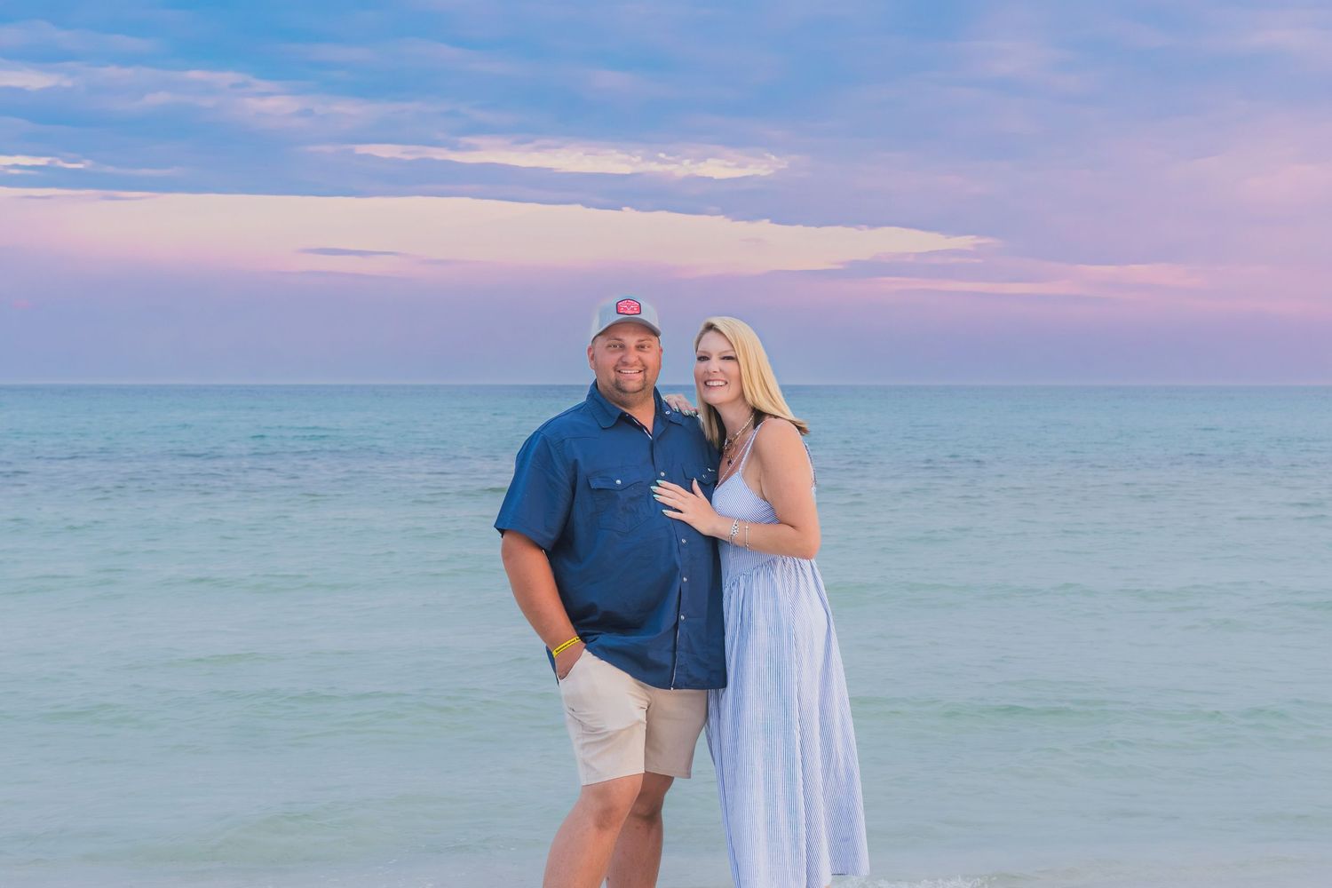 Couple smiling together on a beach at sunset with pastel pink and blue sky over calm turquoise ocean waters.
