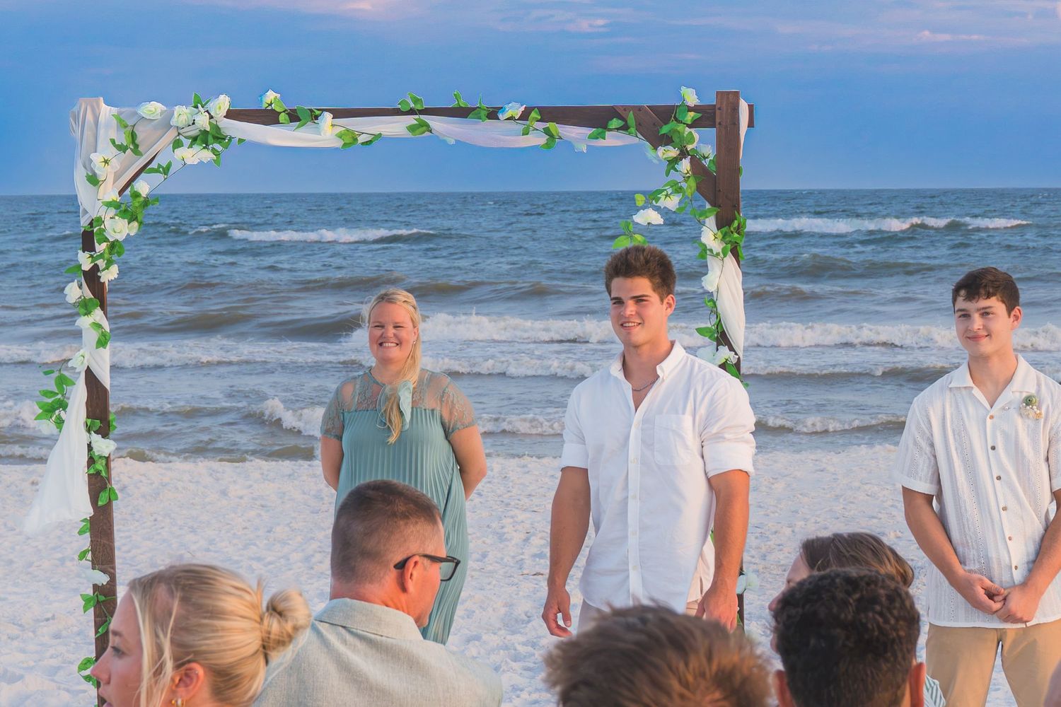 Beach wedding ceremony under floral arch with groom and groomsmen at sunset by the ocean.