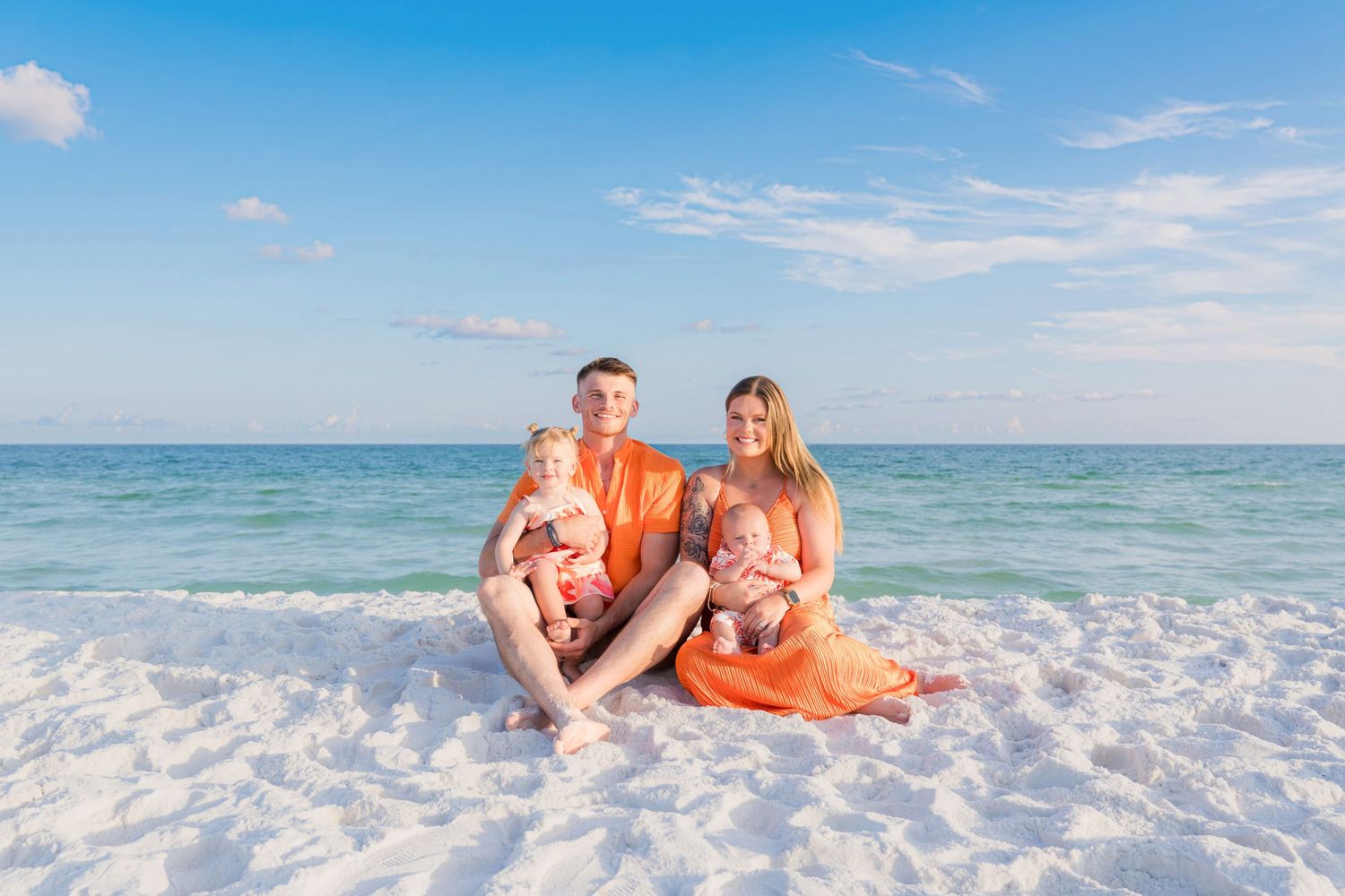 Family of four in matching orange outfits sitting on white sand beach with clear turquoise ocean behind them.