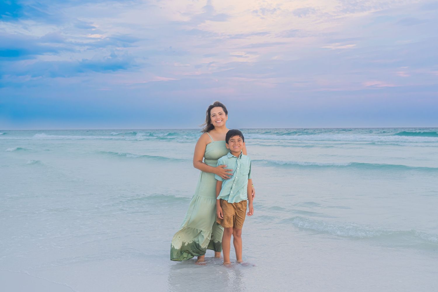 A mother and son stand together on a white sand beach at dusk, wearing coordinating green and tan outfits.