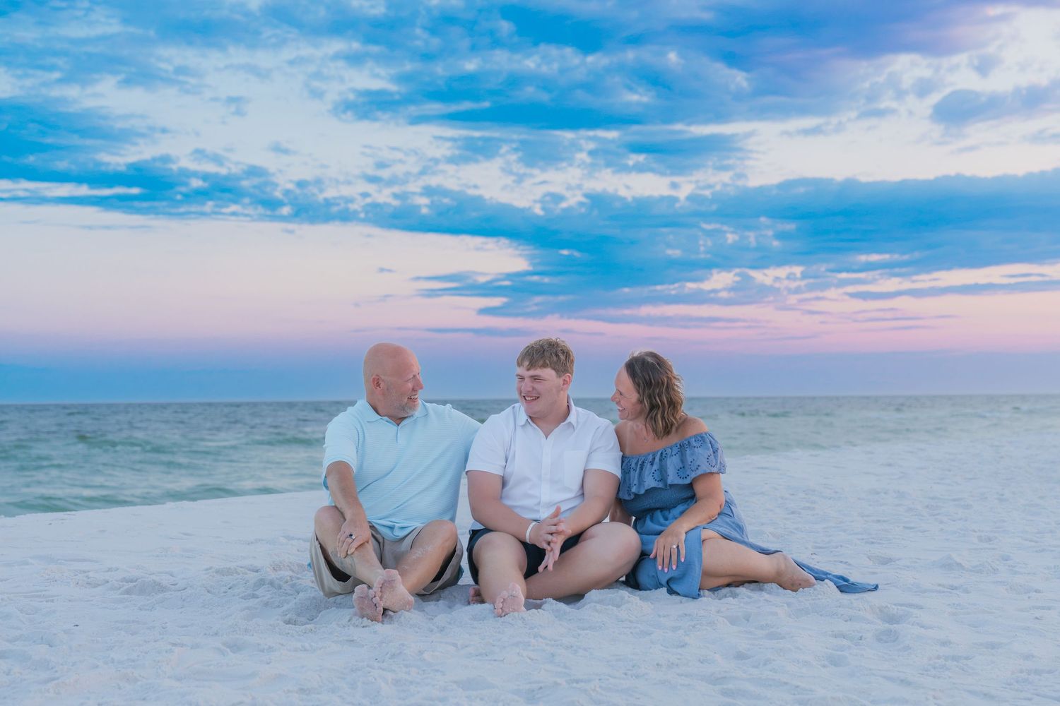 Family of three sitting on white sand beach at sunset with pastel pink and blue sky in background.
