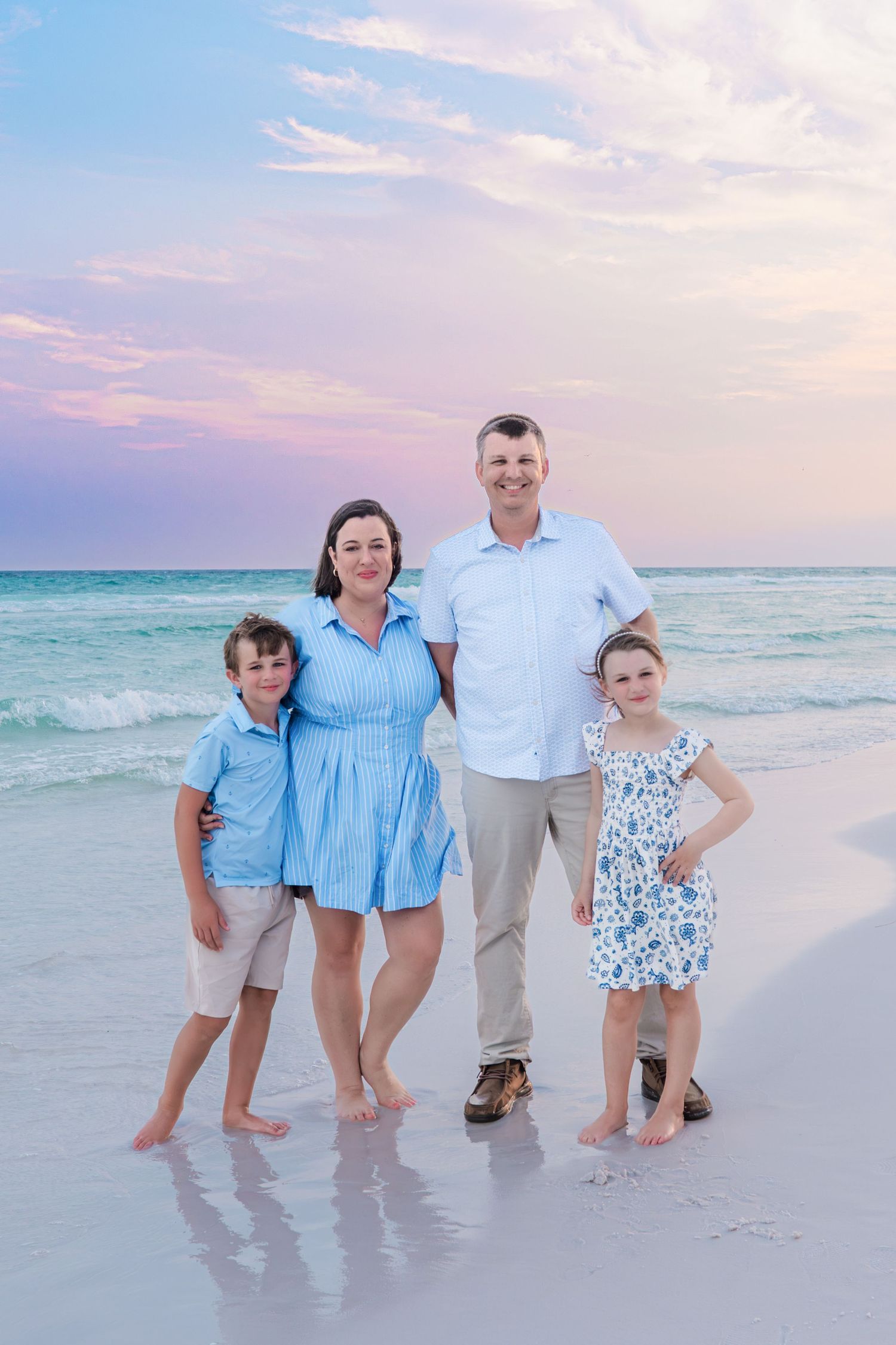 Family of four posing on a sandy beach at sunset, wearing coordinating blue outfits with ocean waves in the background.