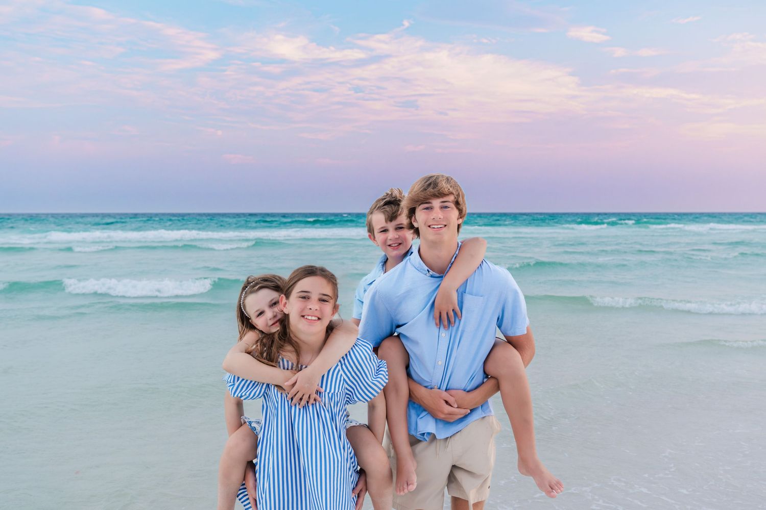 Family of four smiling on a beach at sunset, two kids piggyback riding on teens in blue outfits.