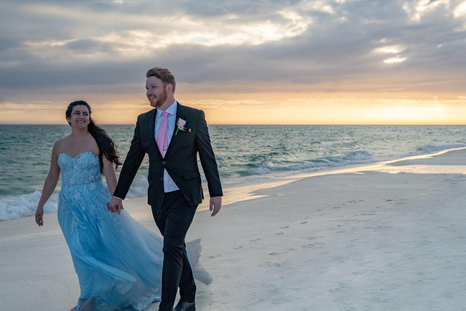 Couple in wedding attire walking hand-in-hand on a beach at sunset, bride in blue gown.