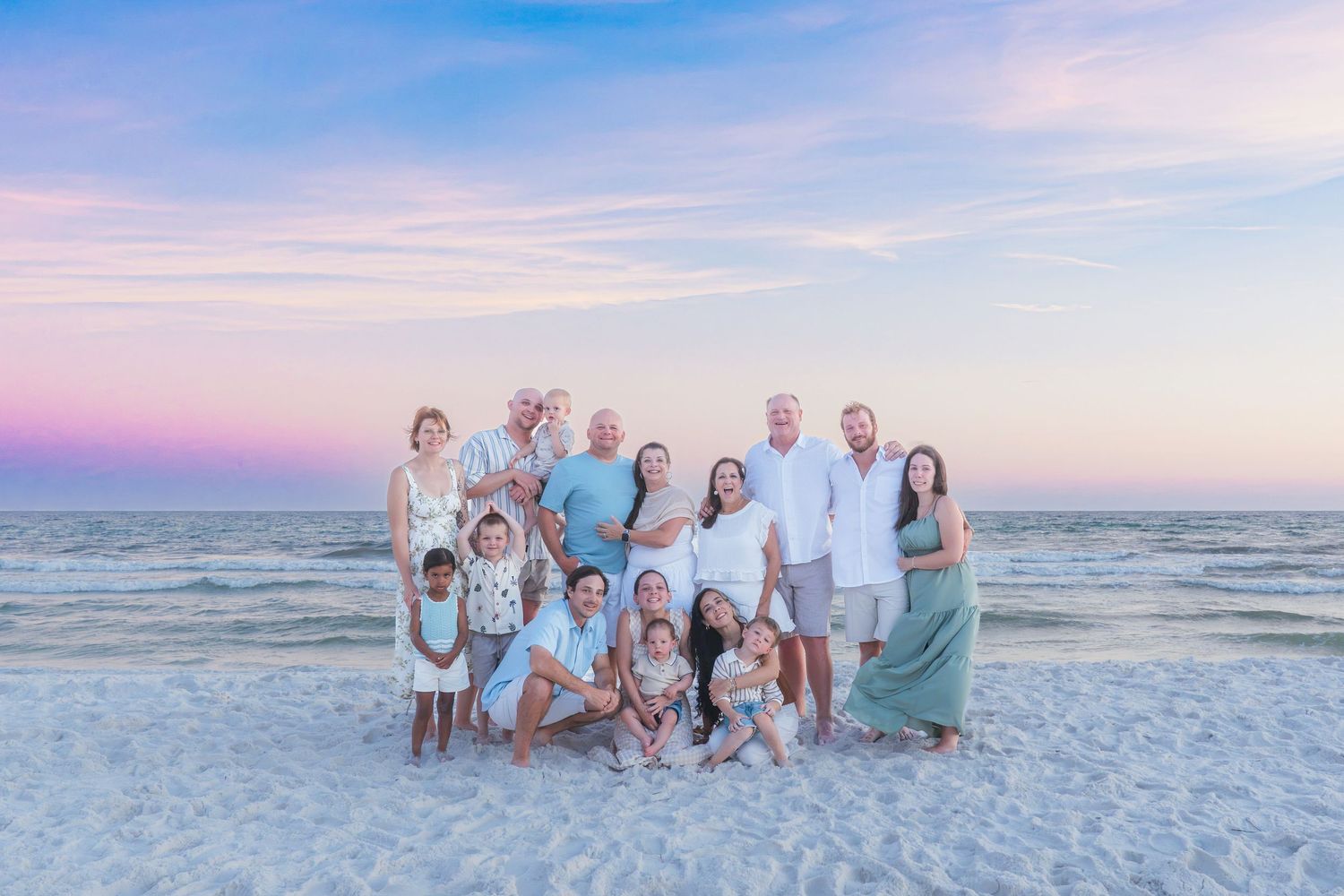 Large family group portrait on a sandy beach at sunset with pink and blue sky in the background.
