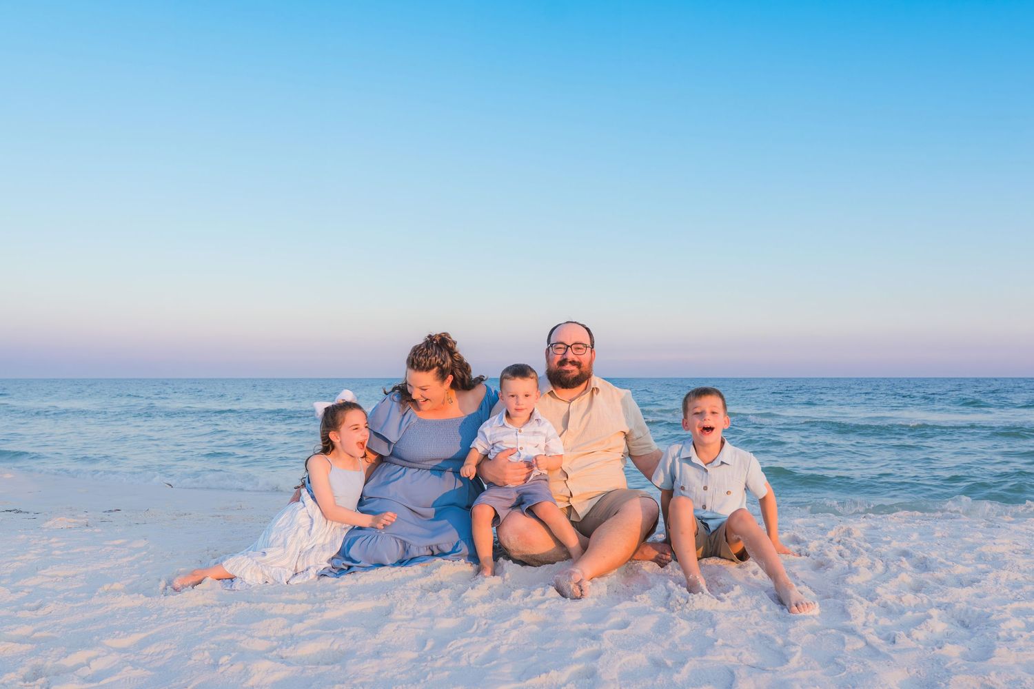 Family of five sitting on white sandy beach at sunset with calm ocean waves in the background.
