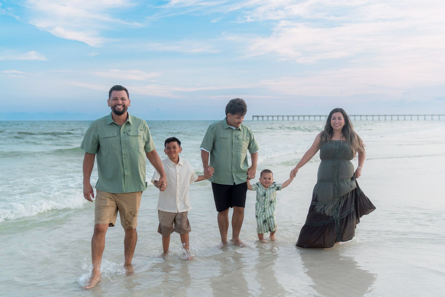 Family of five posing on a beach shoreline at sunset, wearing coordinating green outfits with ocean waves and a pier in background.