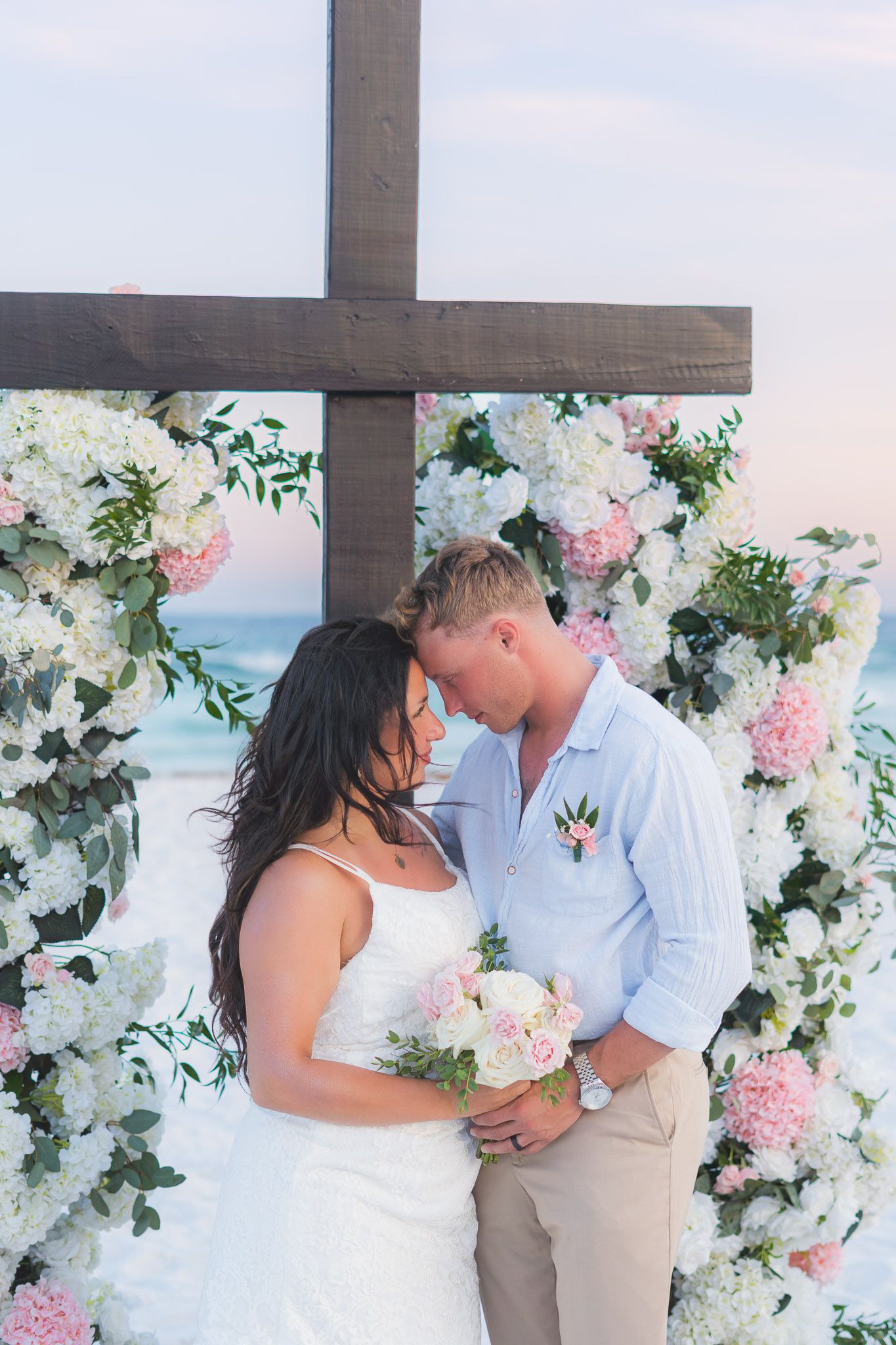 Bride and groom share a romantic moment under a floral arch with a wooden cross at a beach wedding ceremony.