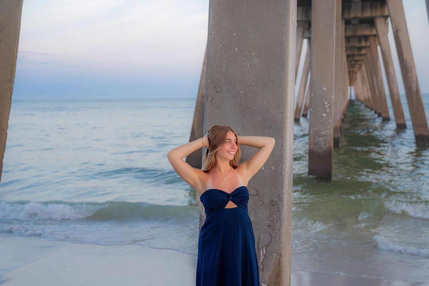 Woman in navy blue gown posing gracefully under a beach pier with ocean waves in the background.