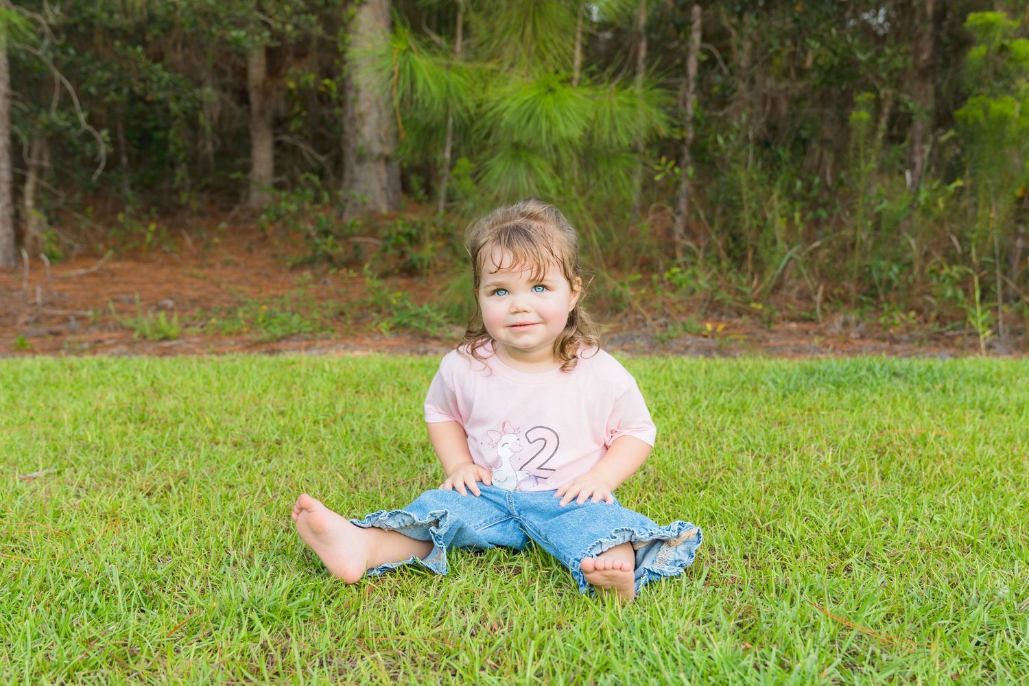 Toddler girl in pink shirt and jeans sitting cross-legged on green grass with pine trees in background.