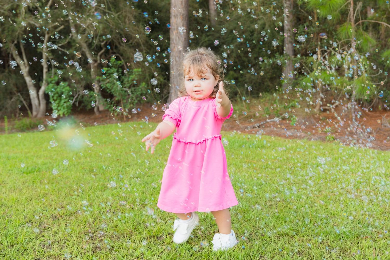 Toddler girl in pink dress playing with bubbles on green grass outdoors.